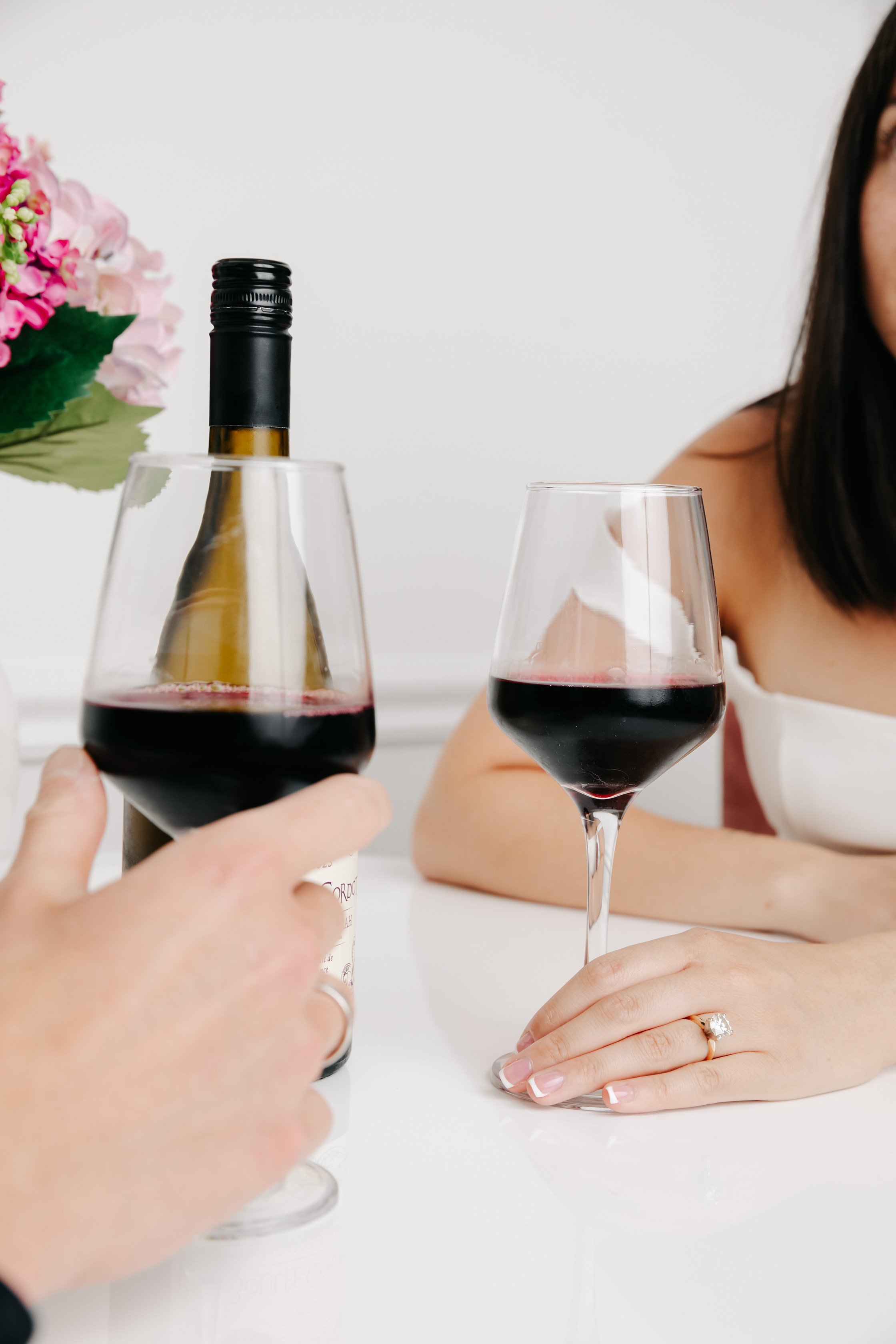 Two women clinking glasses of red wine at a table, with a bottle of wine and a floral arrangement in the background.