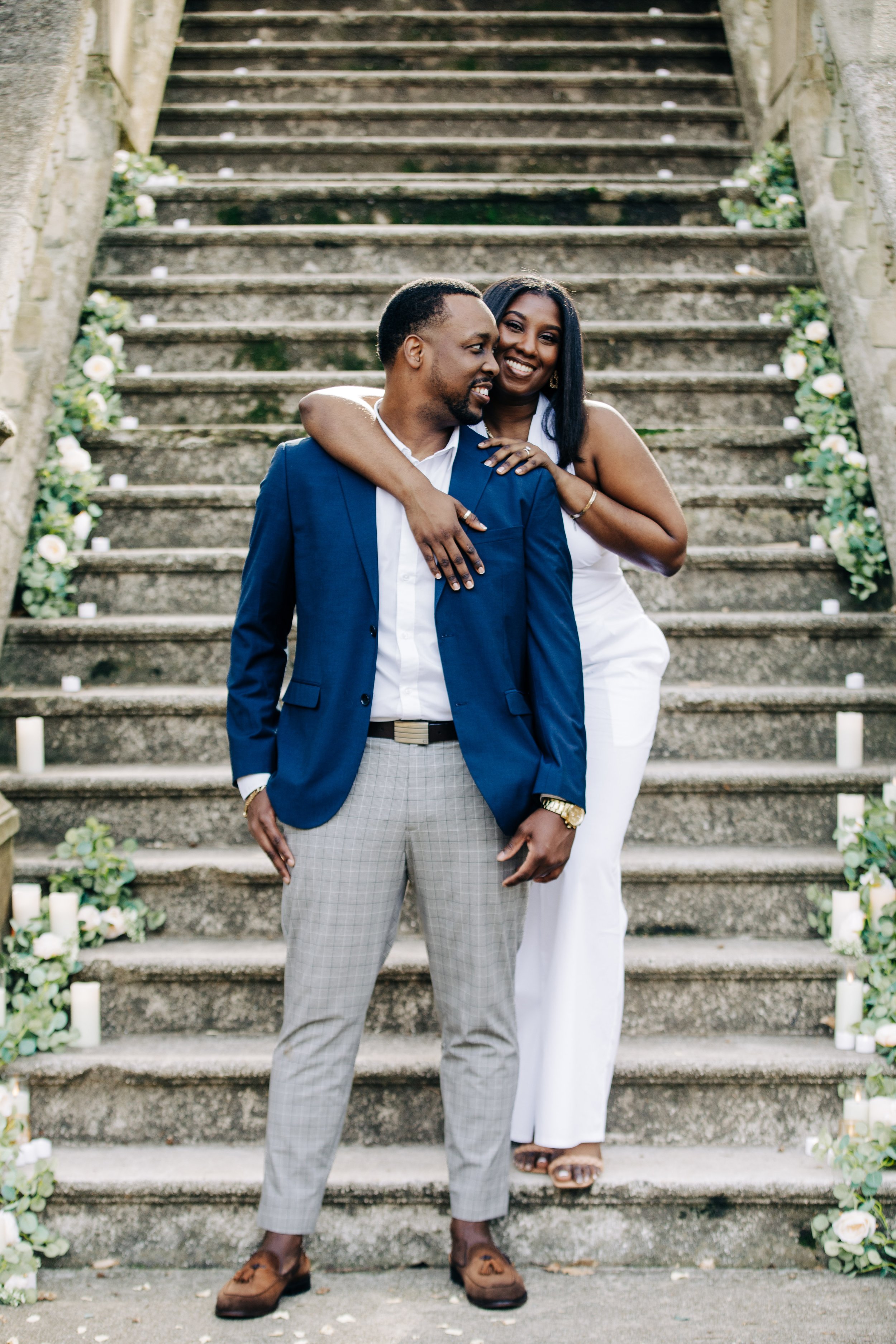 A happy couple standing on outdoor stone stairs decorated with white flowers and candles, smiling at each other, dressed in formal wedding attire.