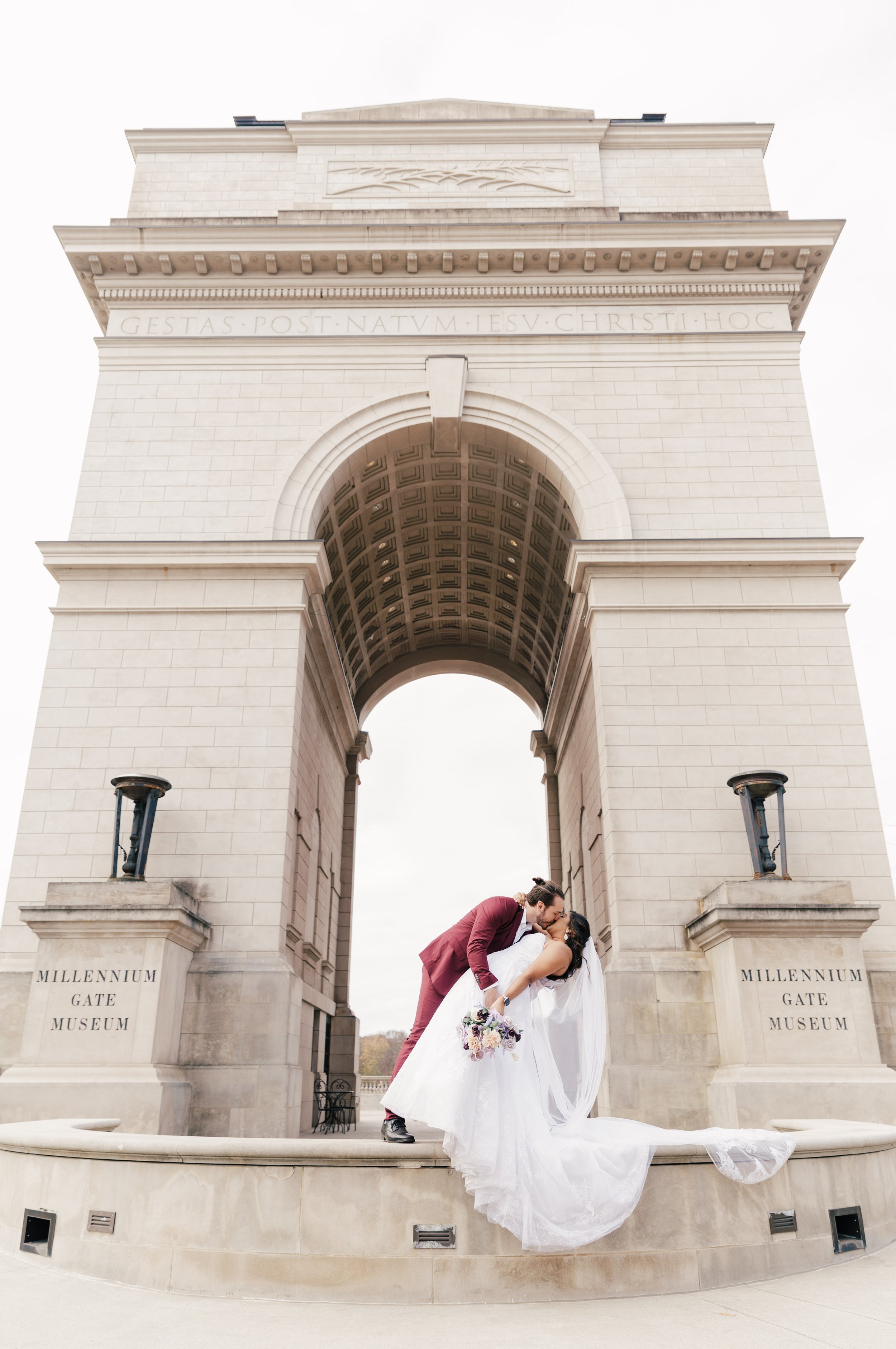 A newlywed couple shares a kiss in front of the Millennium Gate Museum, with the large arch and columns behind them.