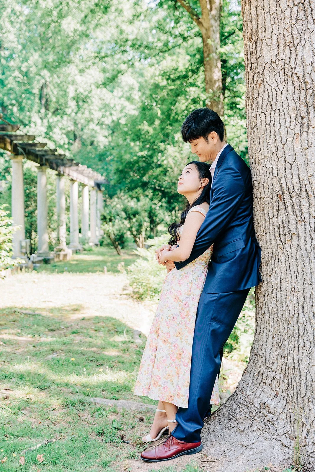 A couple leaning against a tree outdoors, with the man dressed in a blue suit and the woman in a floral dress, sharing an intimate moment.