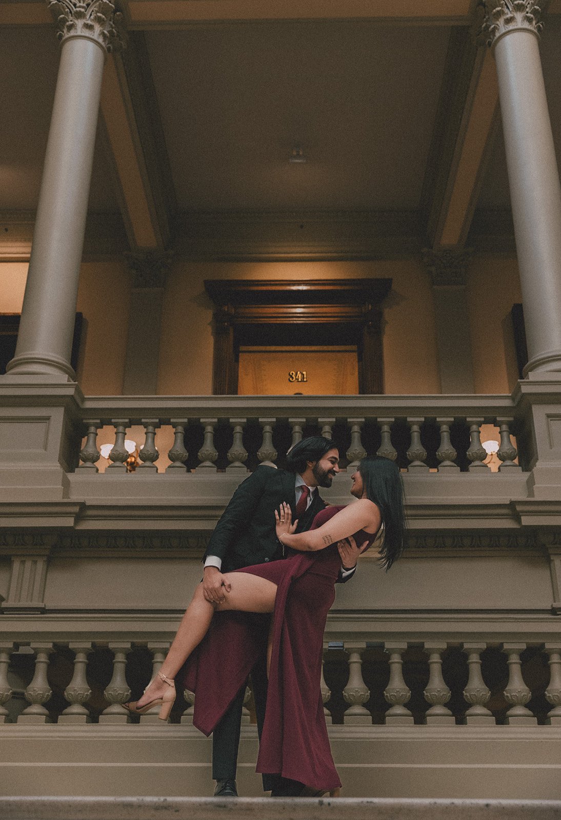 A man in a suit holding a woman in a burgundy dress and heels in his arms, dancing or posing on a staircase in a grand, historic building with ornate wooden details and columns.