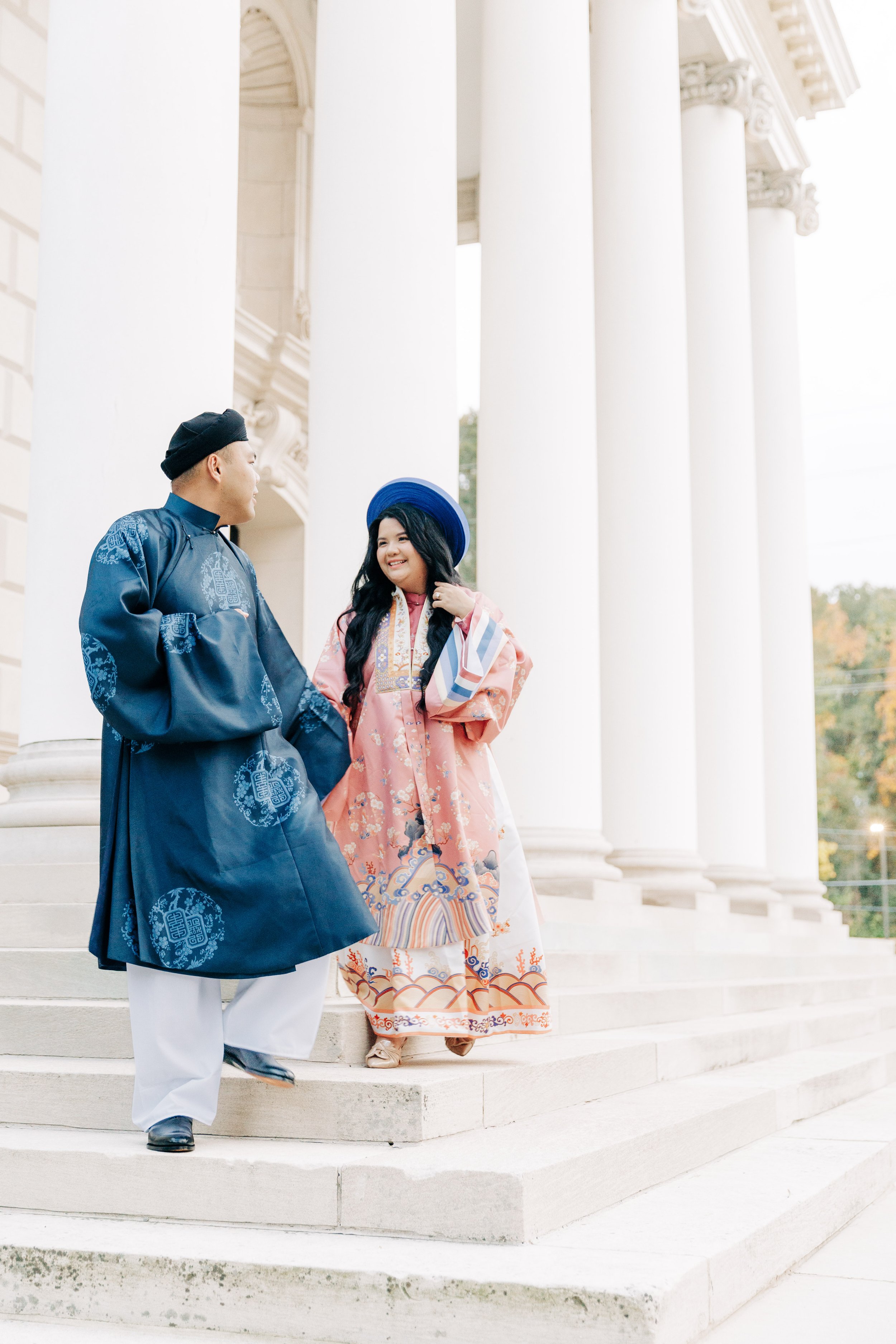 Two people in traditional Asian attire on the steps of a white columned building, smiling and talking to each other.