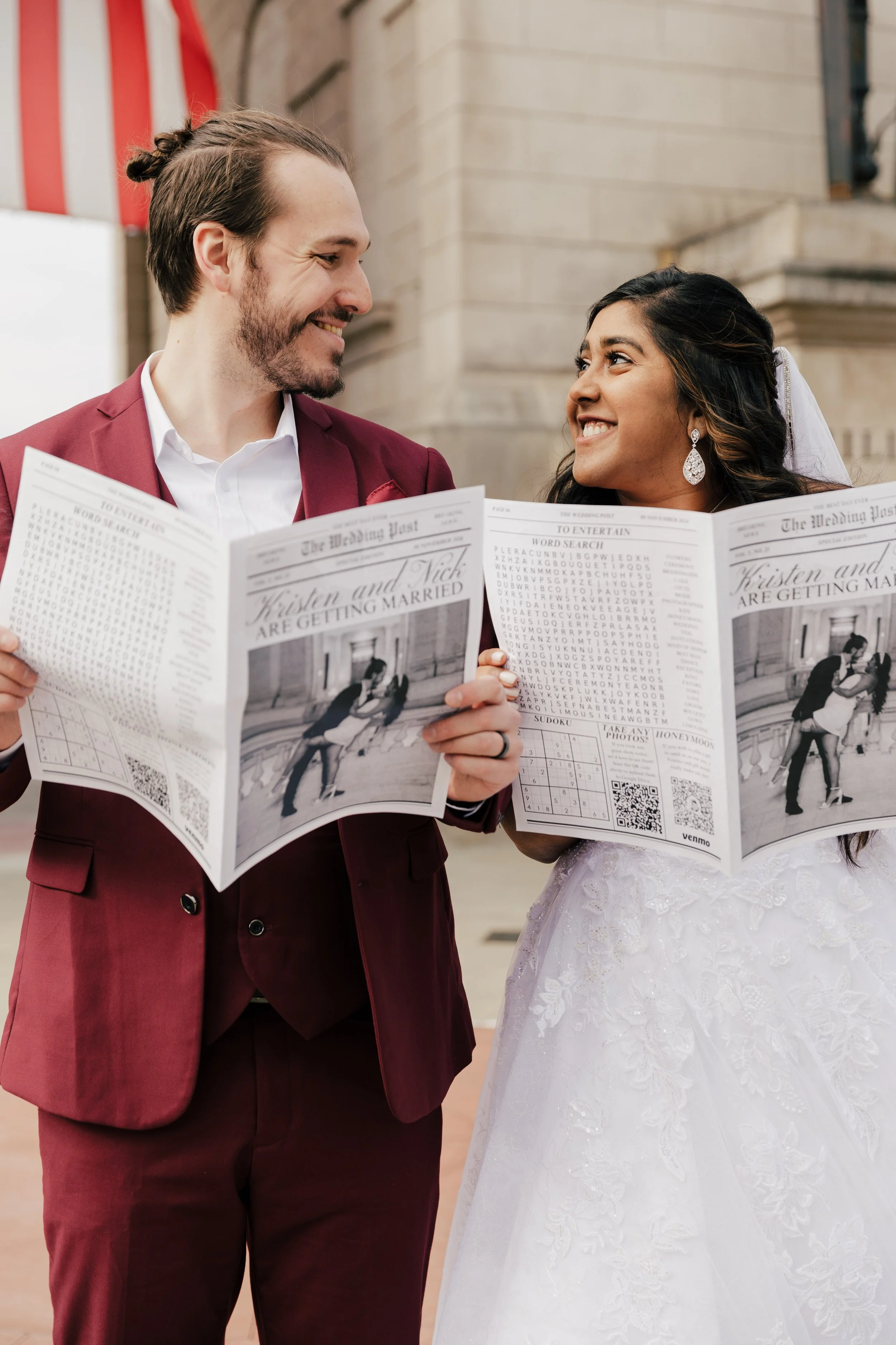 A newlywed couple in wedding attire holding marriage license papers with a happy expression, standing outdoors in front of a stone building.