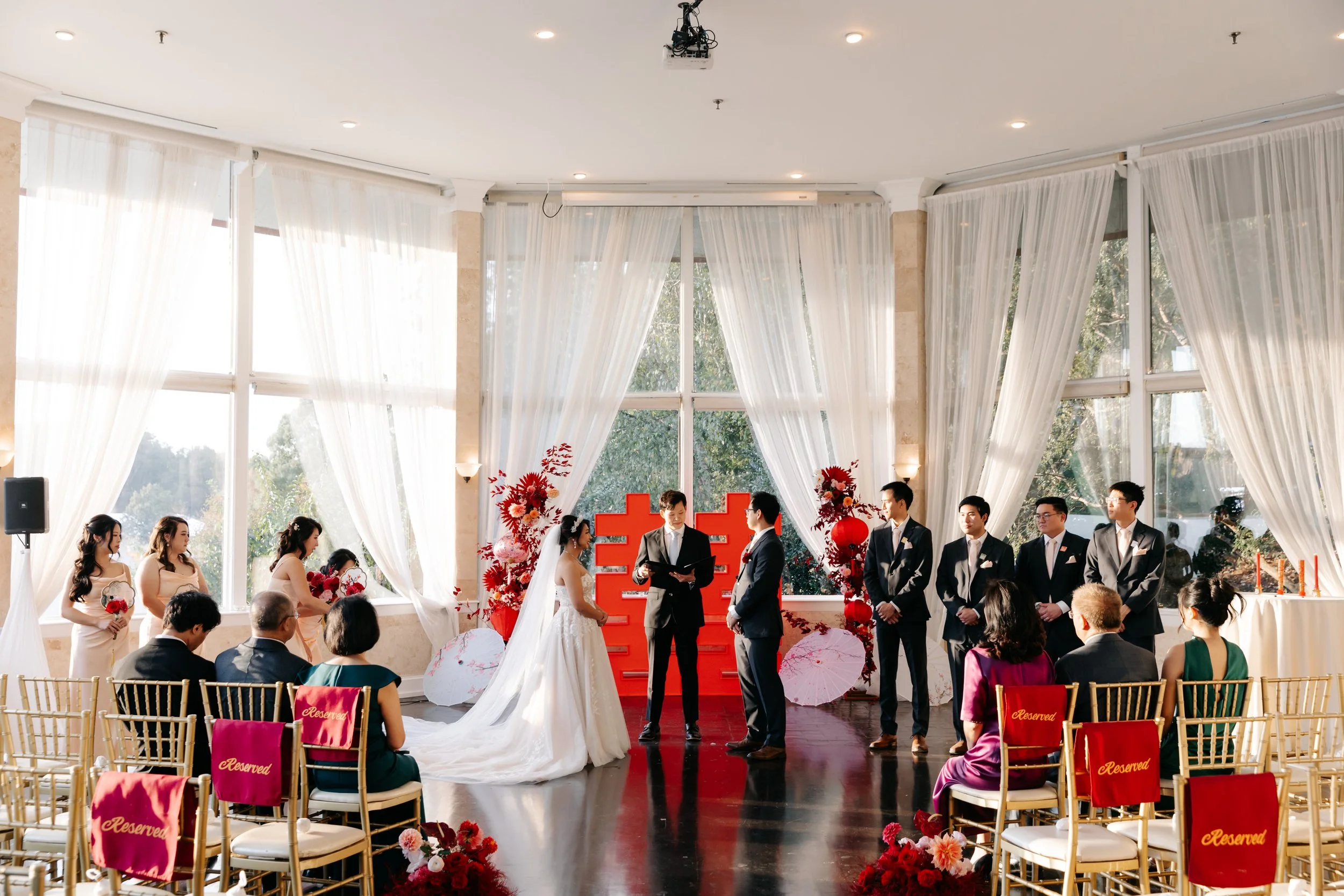 A wedding ceremony with the bride and groom facing each other, officiant reading, in front of decorated background with red flowers and Chinese characters. Bridesmaids and groomsmen are standing on either side, guests seated with reserved signs, and sunlight streaming through large windows.