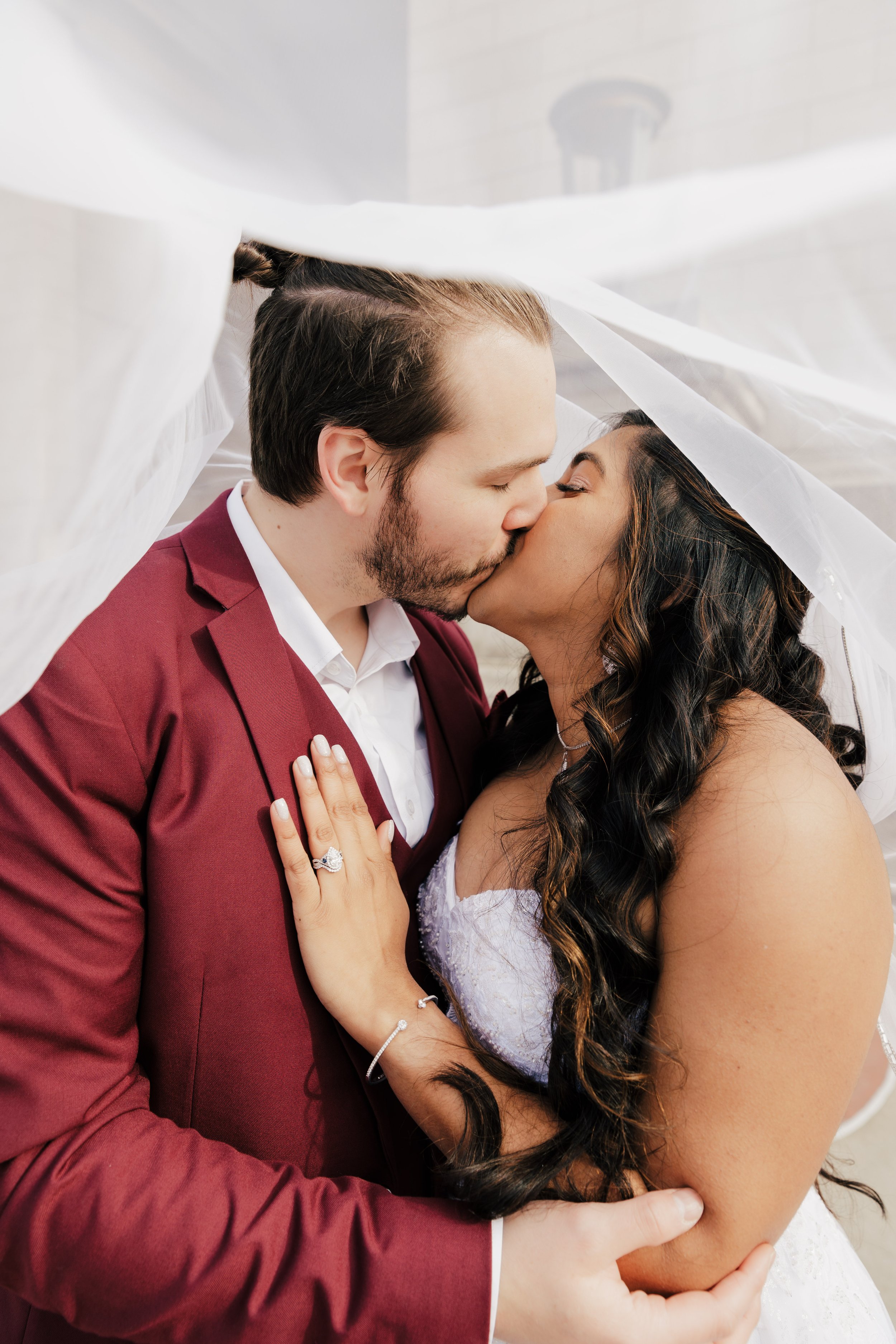 A newlywed couple sharing a kiss under a white veil, with the woman showing an engagement ring.