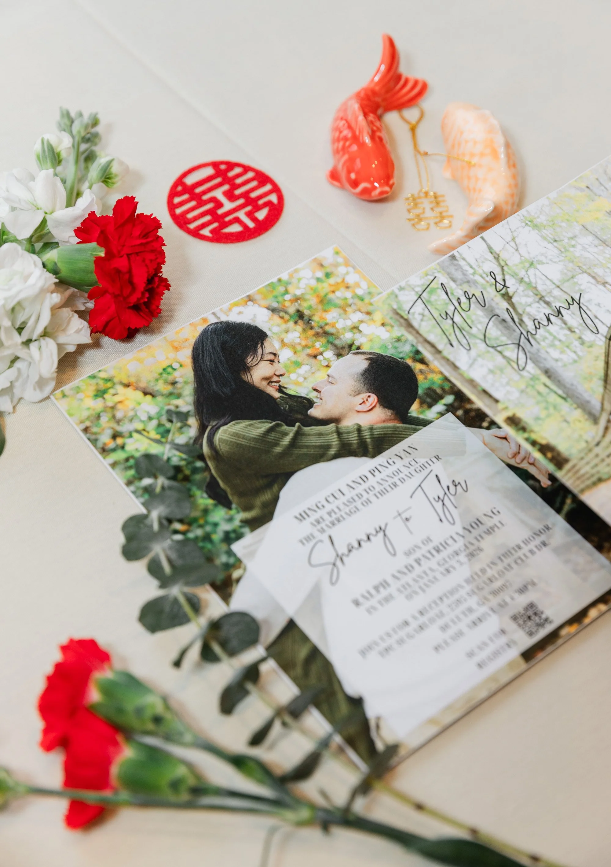 Wedding invitation and photos, with red and white flowers, a red Chinese knot, and gold and coral fish decor on a white tablecloth.