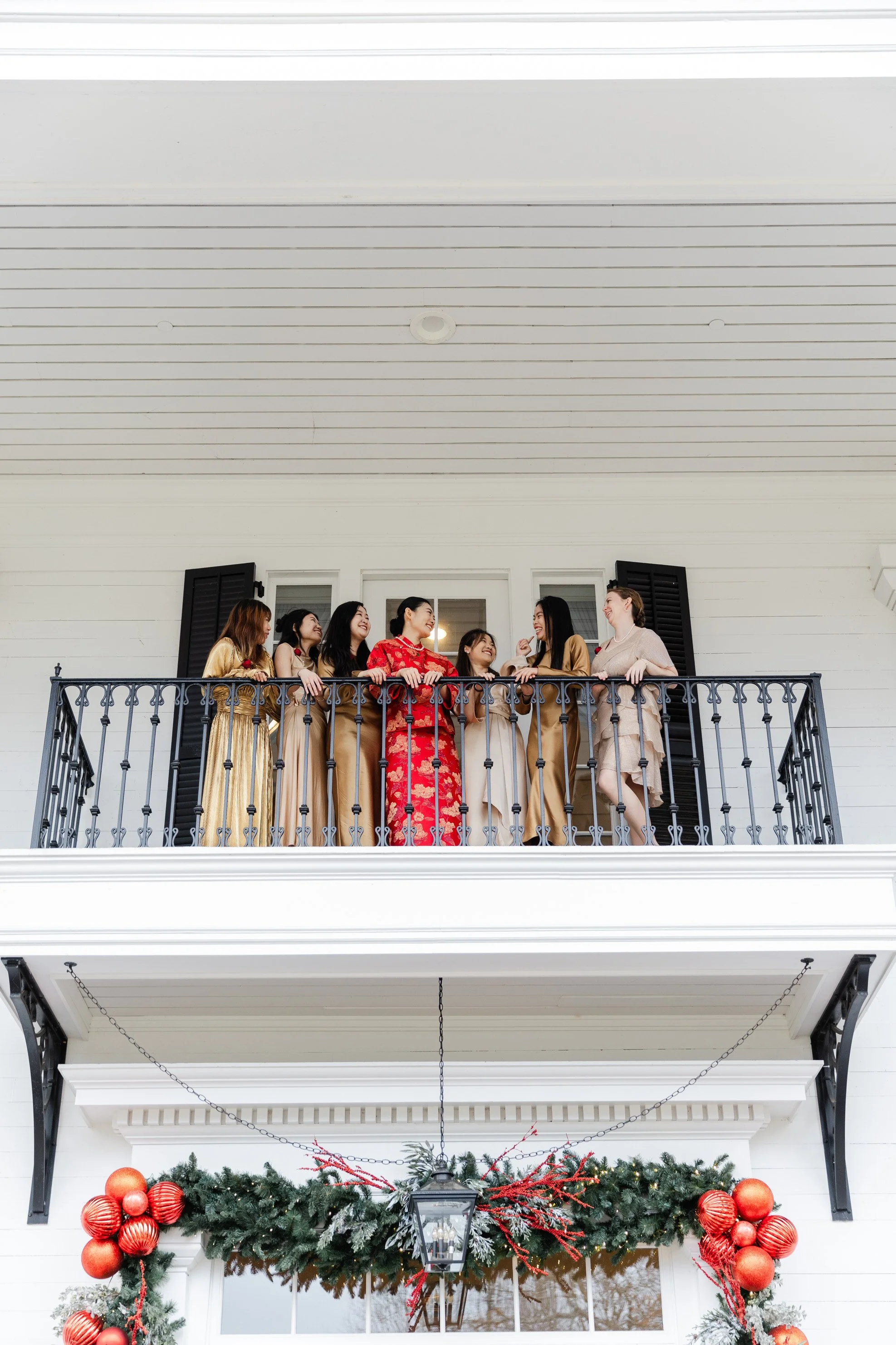 Group of women wearing elegant dresses standing on a house balcony decorated with Christmas ornaments, holiday greenery, and a lantern.