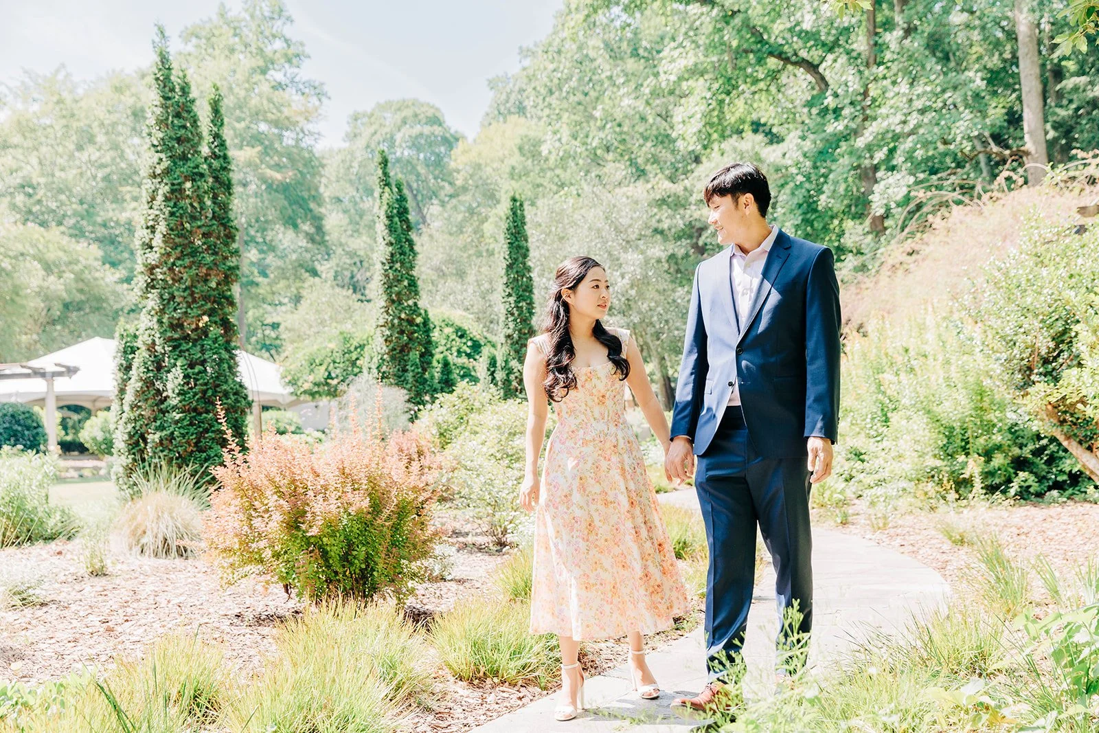 A young woman and a young man walking hand in hand along a garden pathway on a sunny day, surrounded by lush green trees and colorful plants.