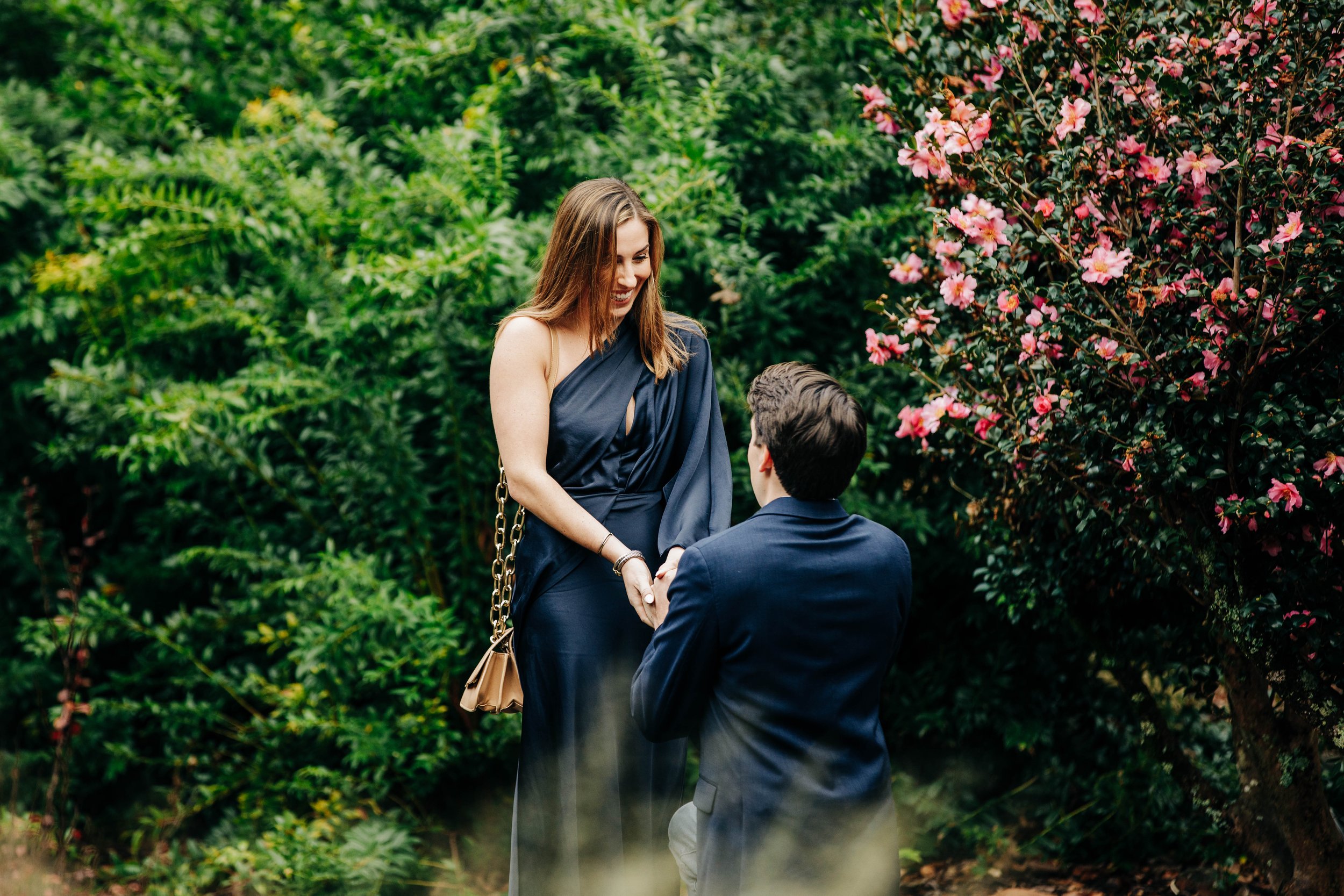 A woman proposing marriage to a man outdoors surrounded by green plants and pink flowers.