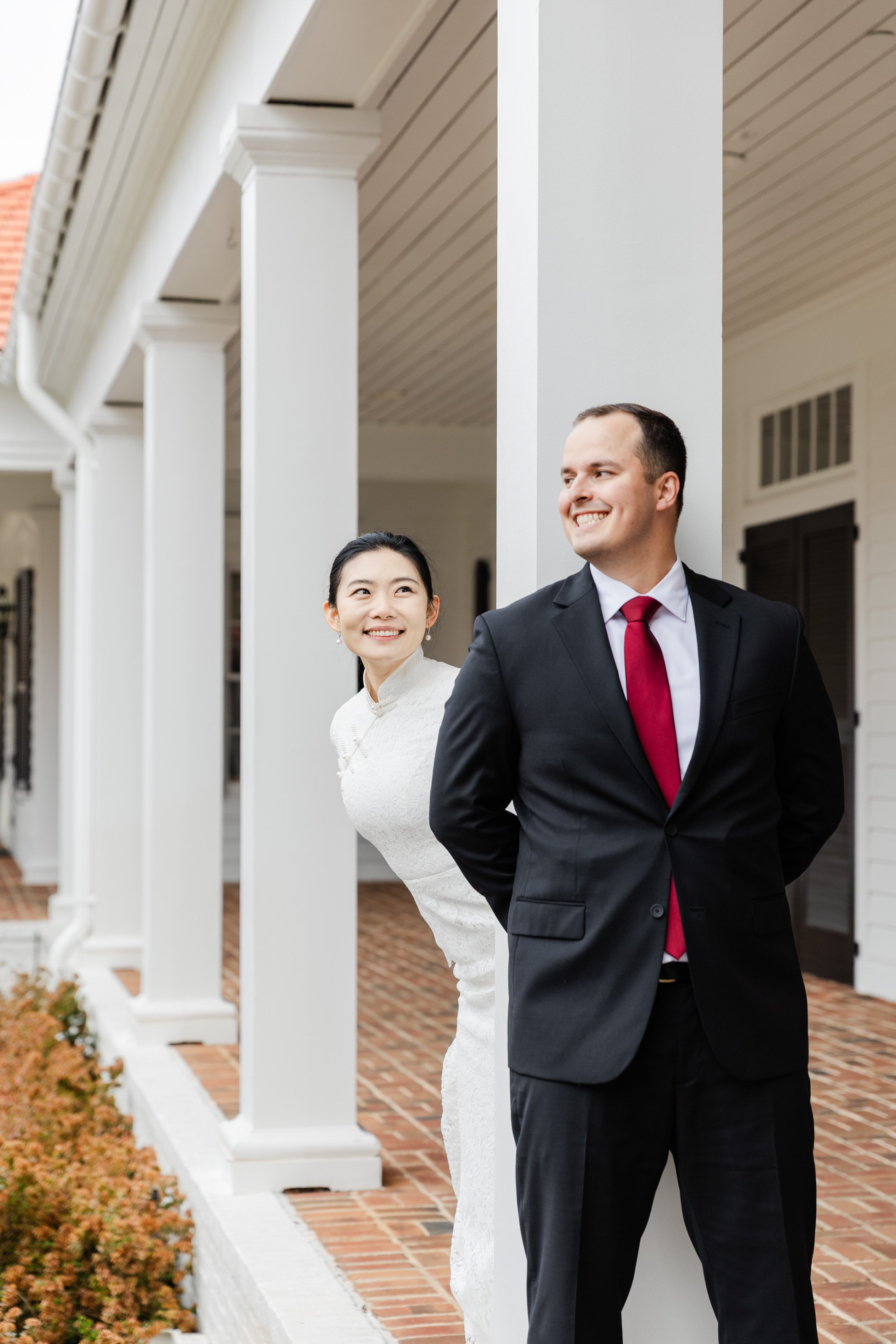 A smiling bride and groom standing outside a white house, with the bride peeking out from behind a column and the groom looking away with his hands behind his back.