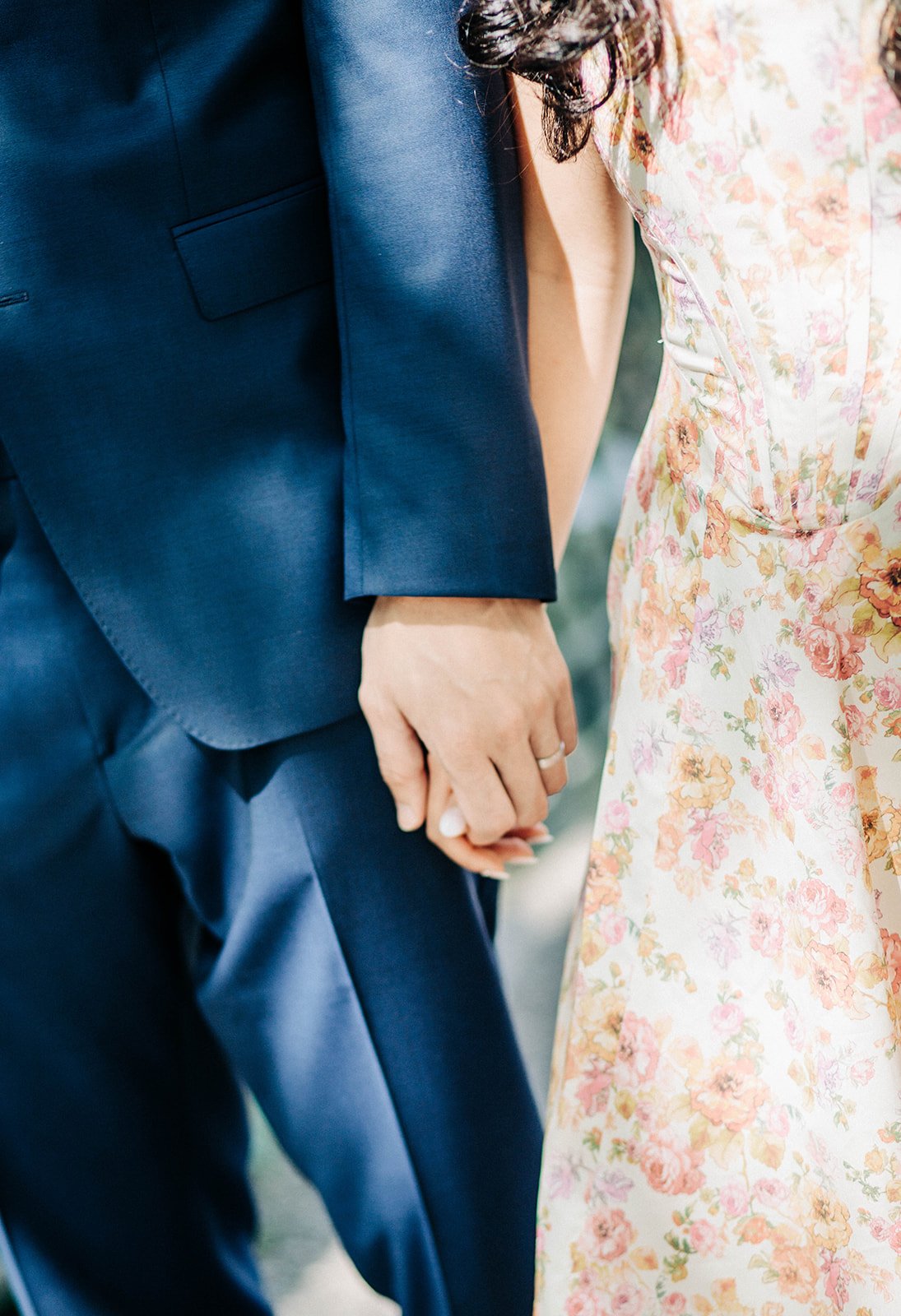 Close-up of a man and woman holding hands, dressed in formal attire, with the man in a navy suit and the woman in a floral dress.