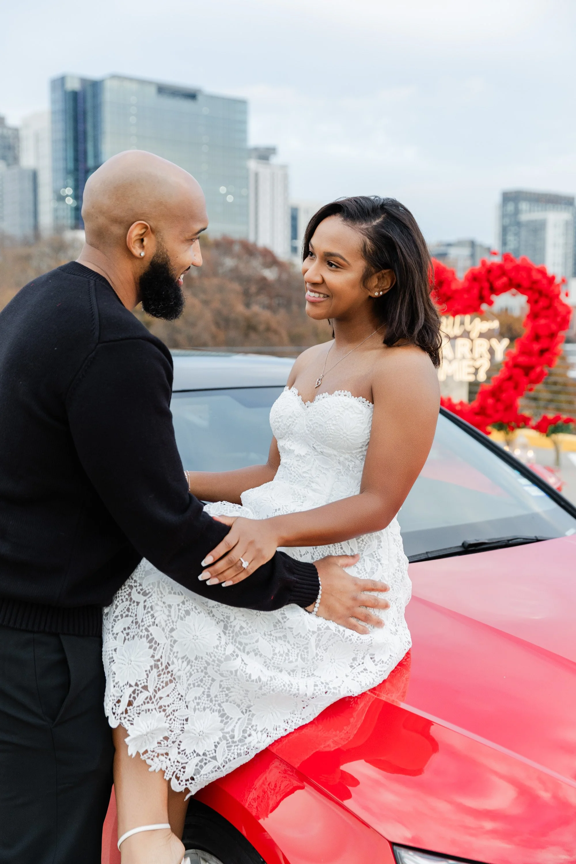 A couple on a wedding day, the bride sitting on a red car, holding hands, smiling, with a city skyline in the background and a red heart-shaped decoration with flowers.