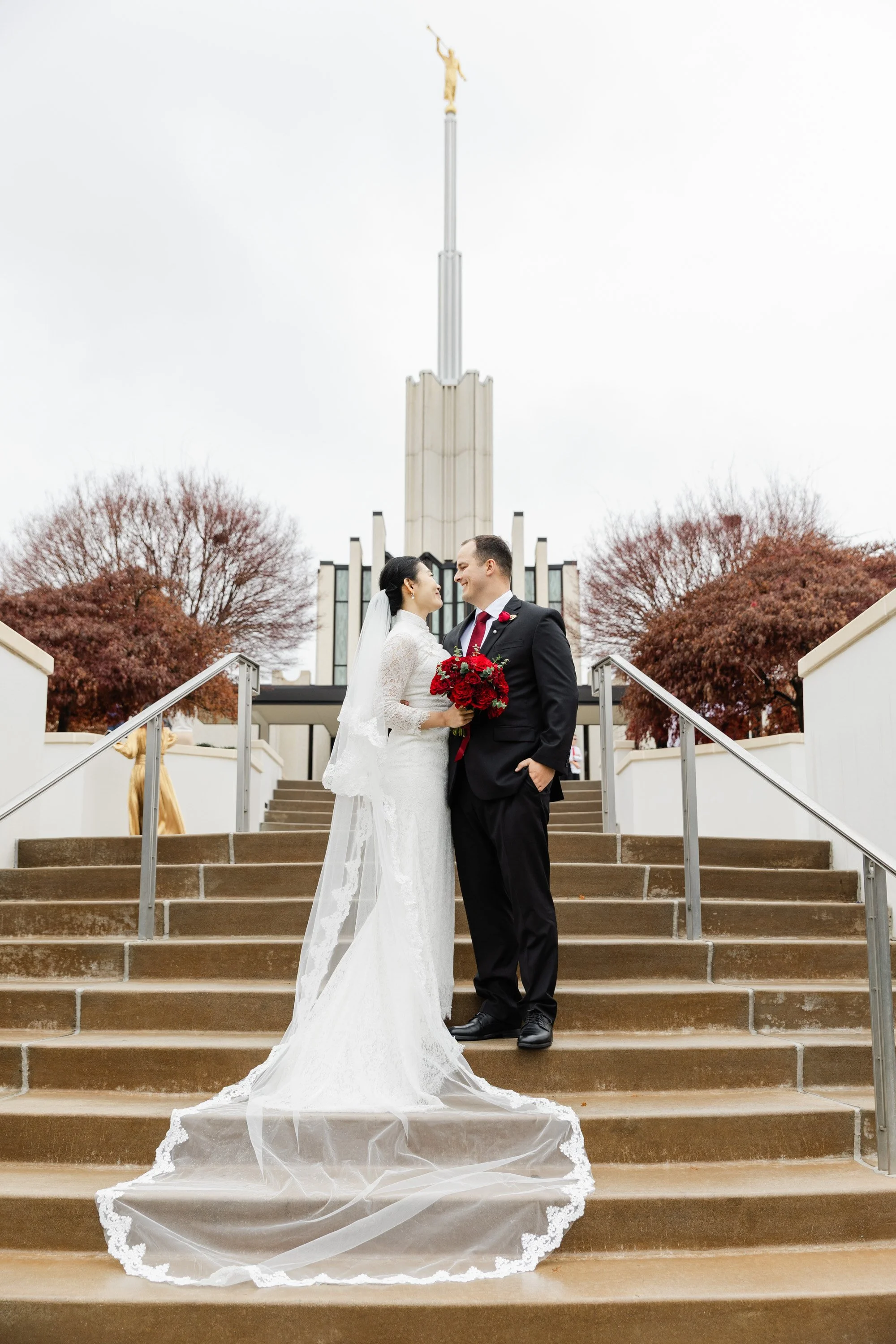 Bride and groom standing on stairs outside, facing each other, with the bride holding a bouquet of red flowers. The bride is in a white lace wedding dress with a long veil, and the groom is in a black suit with a red tie and white shirt. Behind them 
