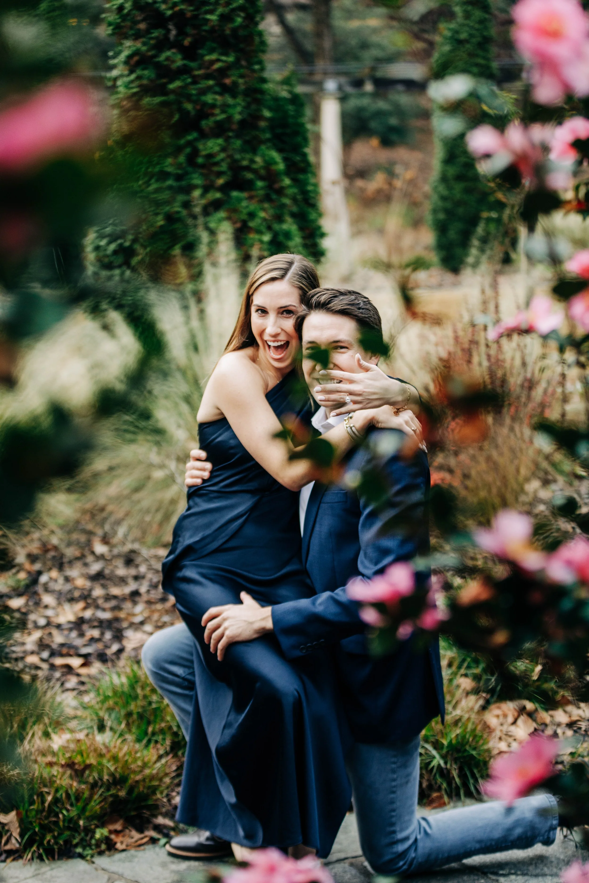 A happy couple in formal attire, with the woman sitting on the man's knee, surrounded by pink flowers and greenery in a garden setting.