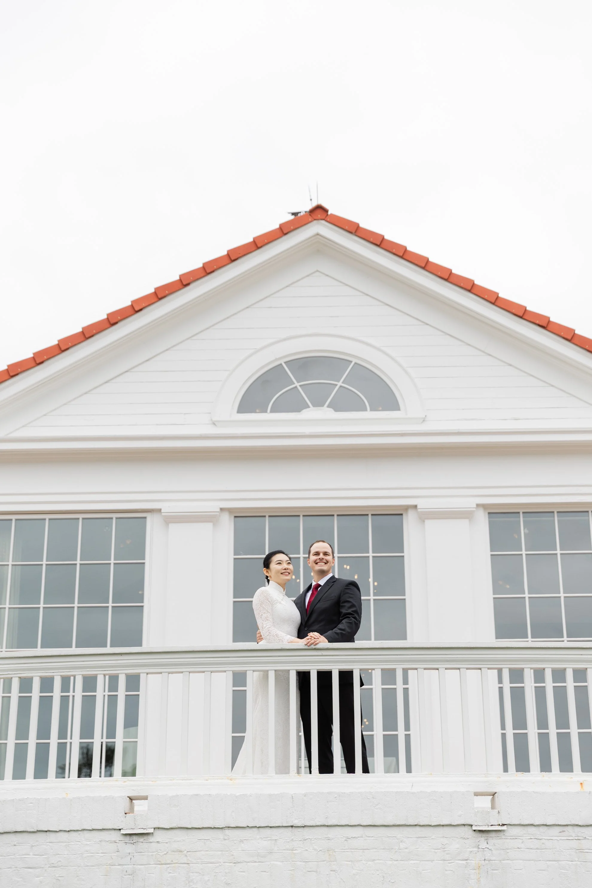 A newlywed couple standing on a white balcony in front of a white building with large windows and a triangular roof with red tiles, smiling and holding hands.