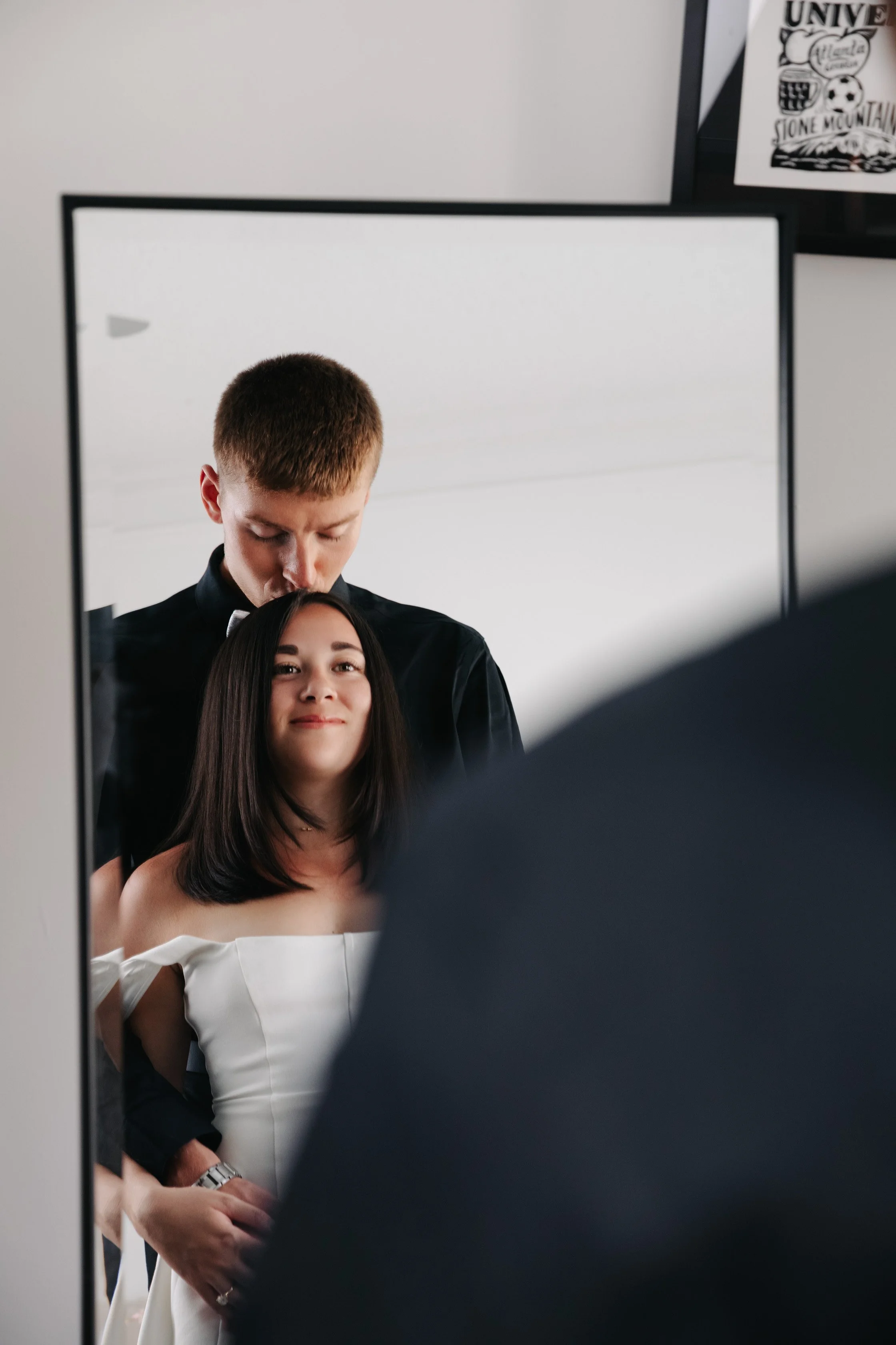A woman with shoulder-length dark hair smiling at her reflection in a mirror, with a man behind her looking down, in a room with white walls.
