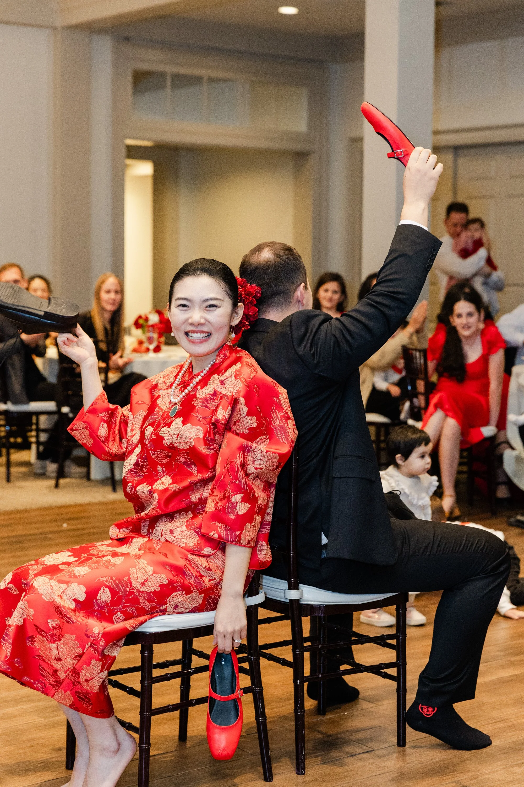 A woman in a red floral dress and a man in a black suit sitting back-to-back, holding shoes and raising one hand each, participating in a game during a celebration with onlookers smiling.