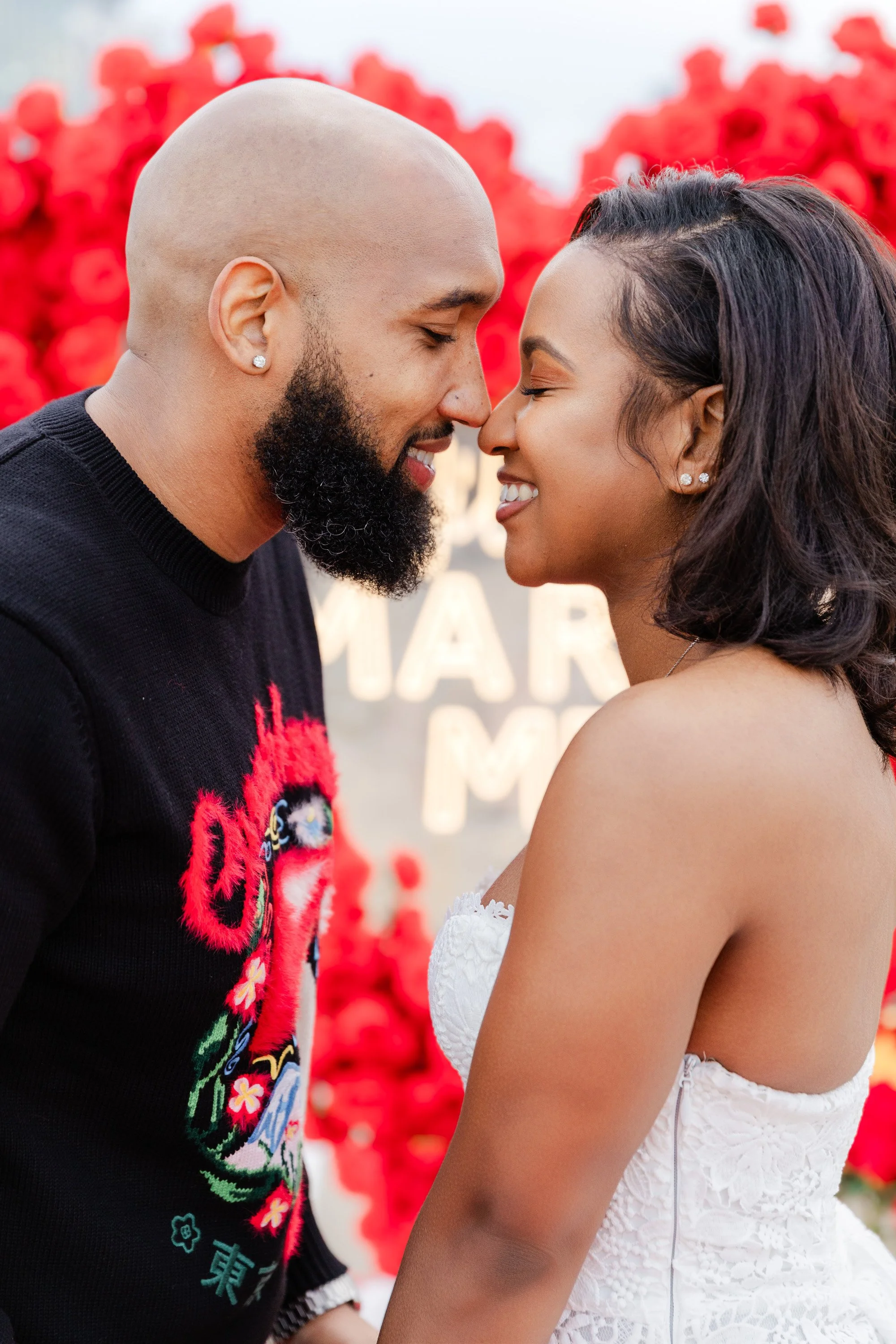 A man and woman face to face, touching foreheads, with happy expressions, at a romantic outdoor setting with red flowers in the background.