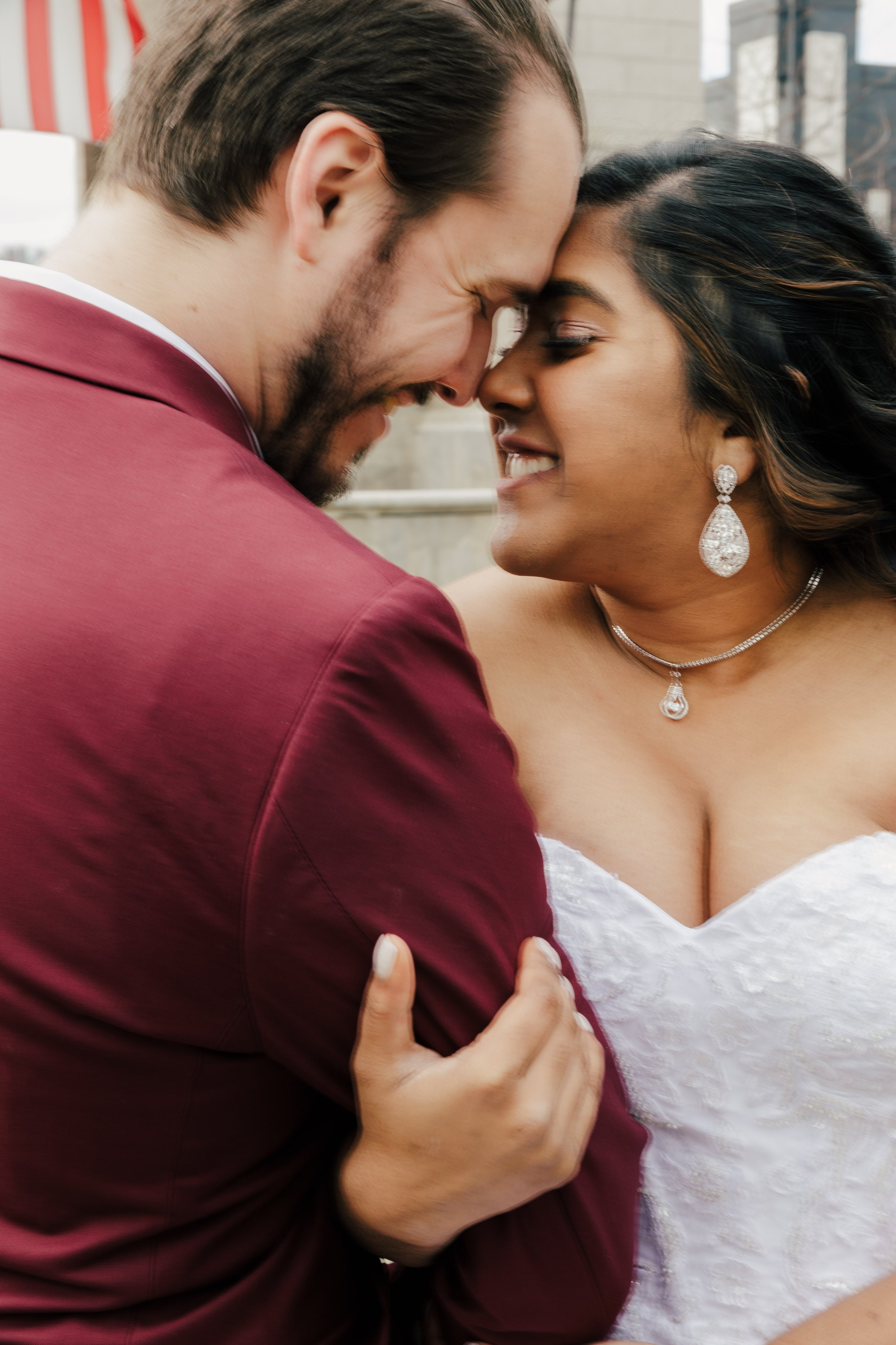 A couple is facing each other with their foreheads touching, smiling at each other. The woman is wearing a white wedding dress and jewelry, while the man is in a burgundy suit.