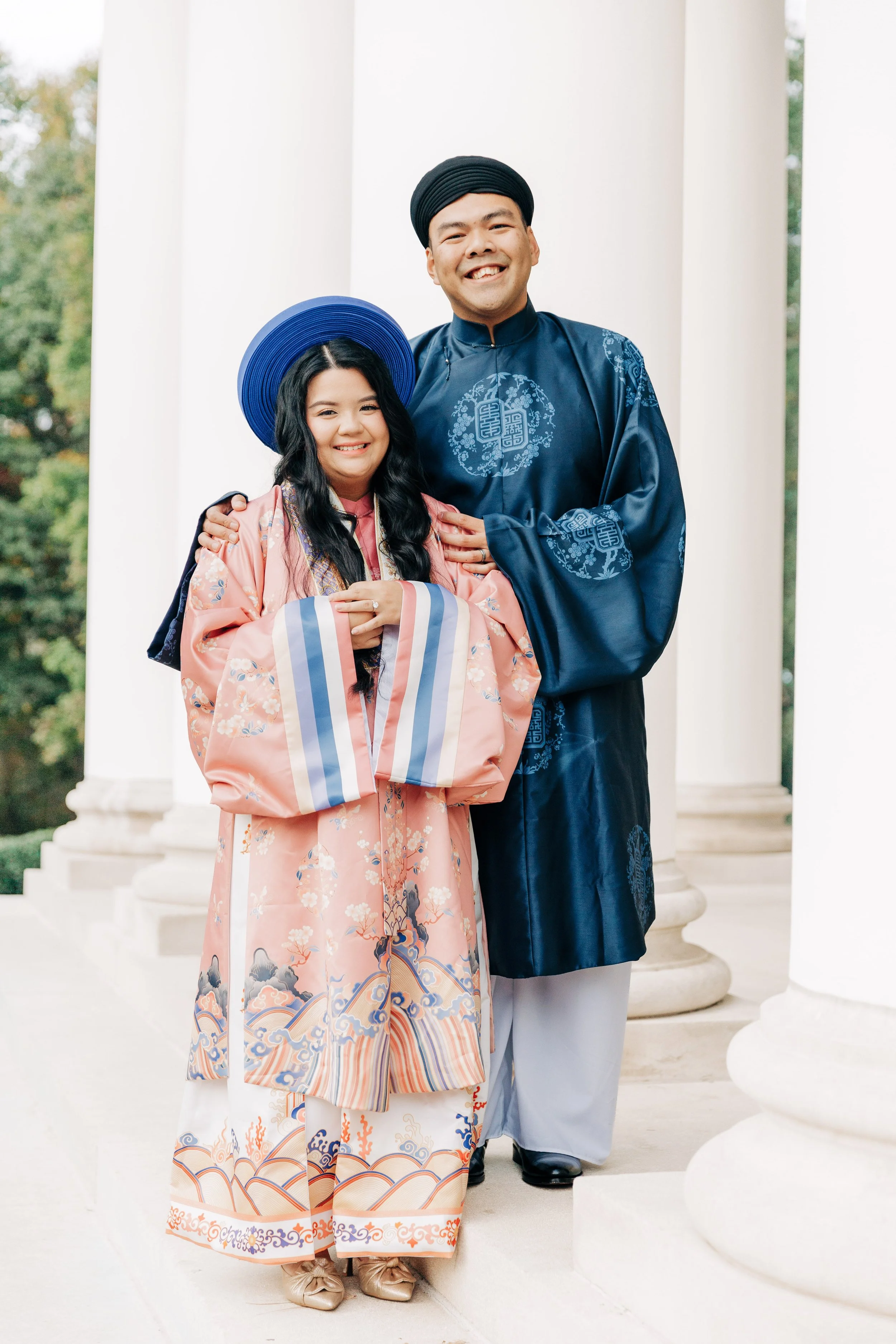 A young woman in a traditional Asian dress and blue hat, and a man in a blue traditional robe and black turban, standing together and smiling in front of white columns outdoors.