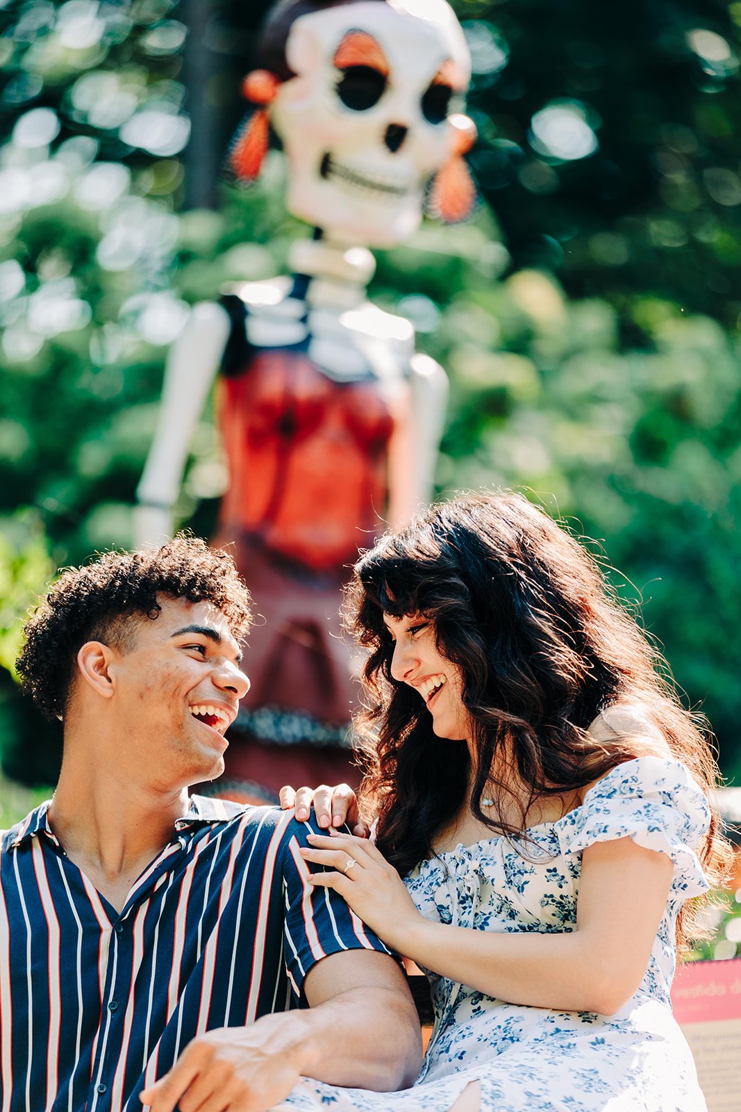 Two people, a man and a woman, smiling and looking at each other outdoors, with a skeleton figure in the background.
