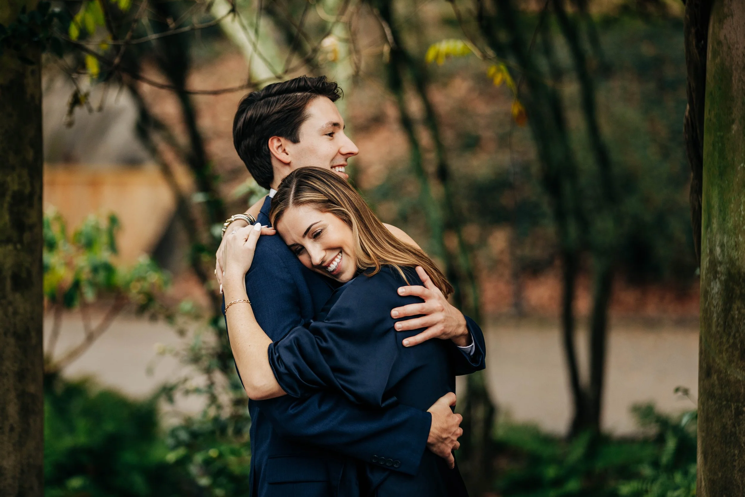 A happy couple in formal attire sharing a hug outdoors in a park with trees and greenery.