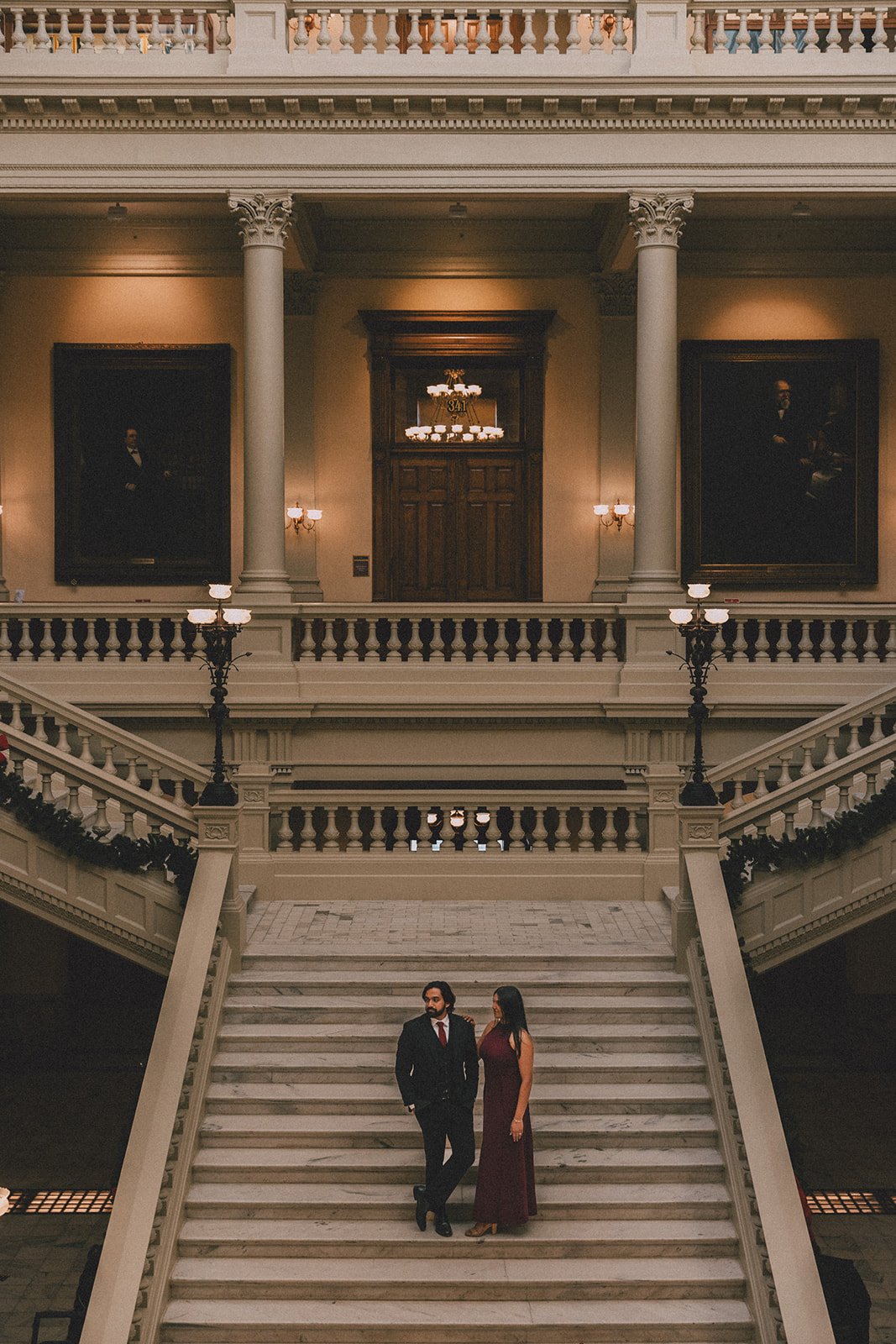 A man in a black suit with a red tie and a woman in a long burgundy dress stand on a grand marble staircase inside a historic building with ornate railings, large portraits on the walls, and warm lighting.