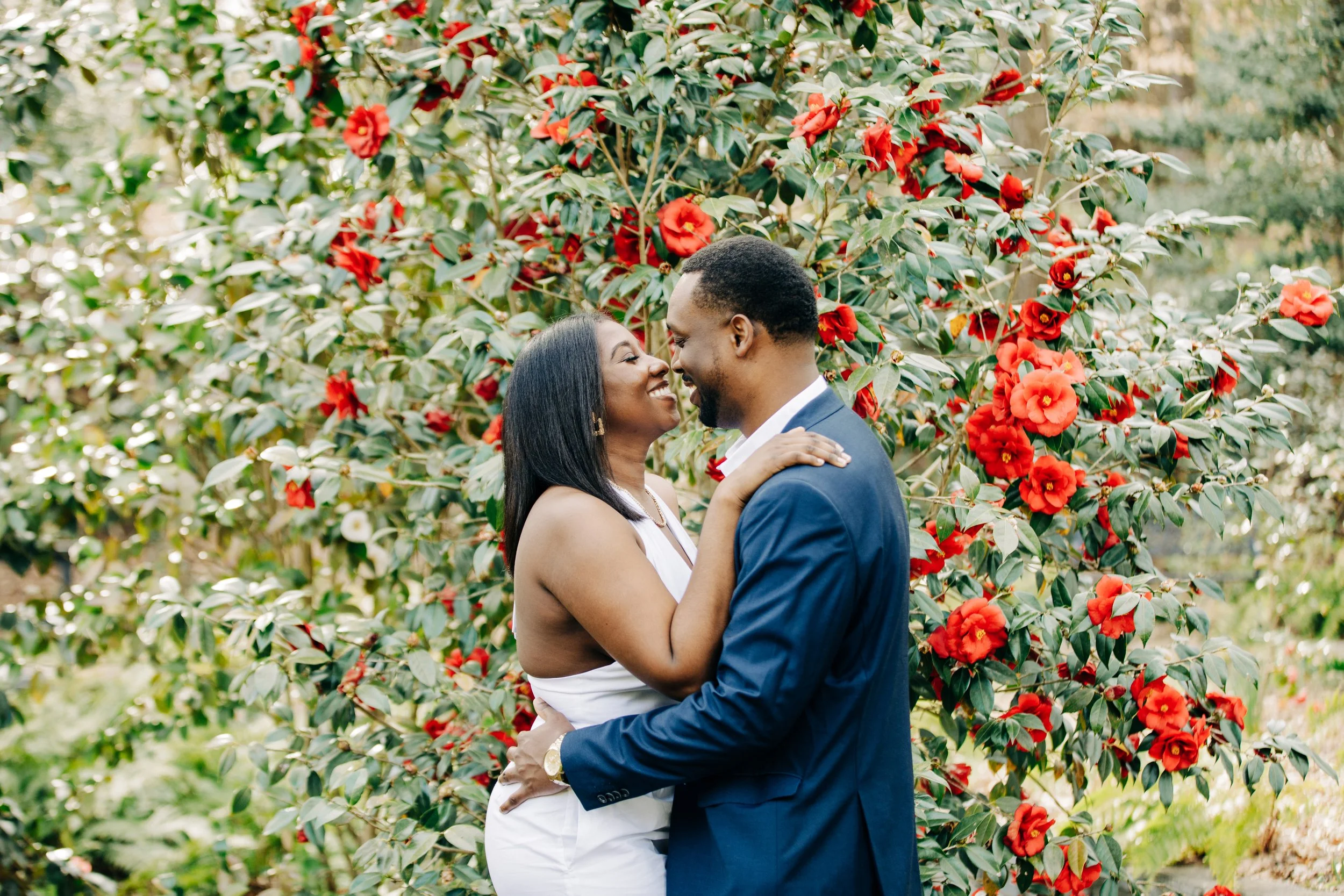 A couple in formal attire sharing a romantic moment in front of a lush flowering bush with red and orange flowers.