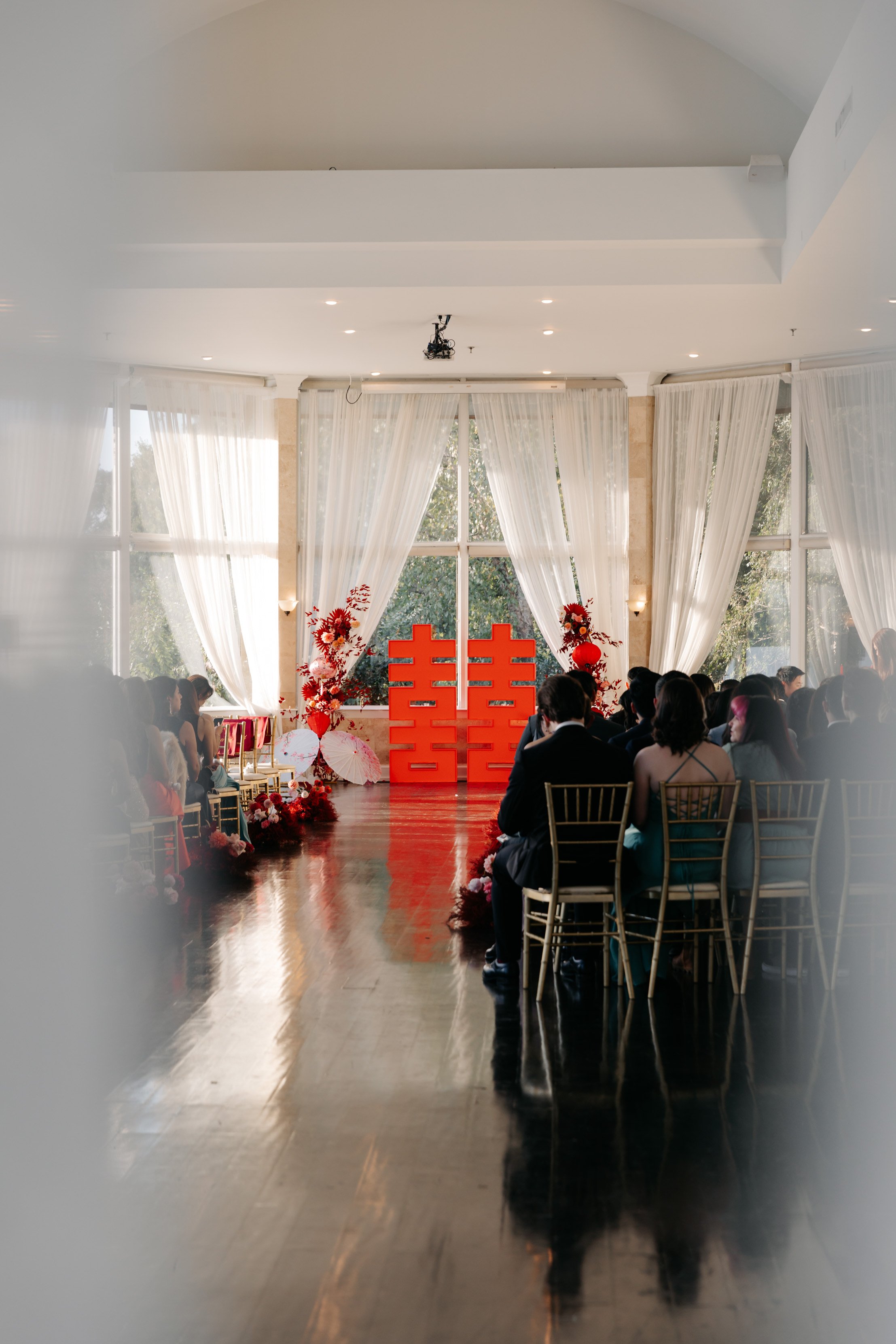 Wedding ceremony setup with large red double happiness symbols on stage, surrounded by red floral arrangements, white curtains, and guests seated on gold chairs.