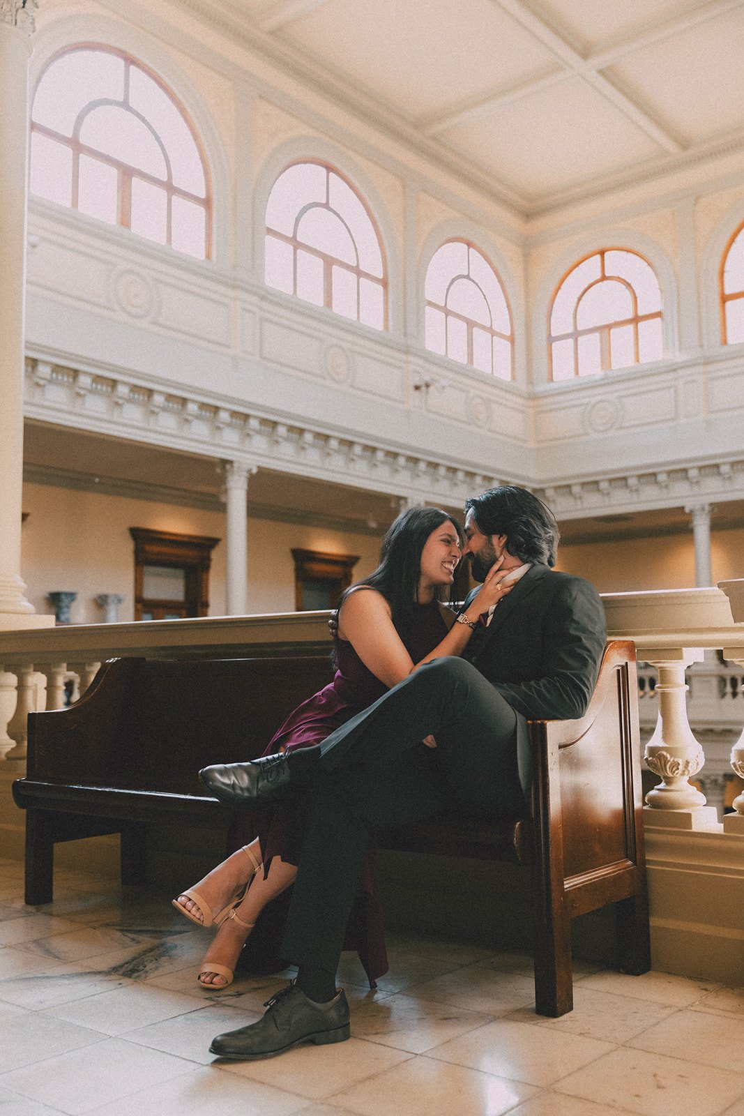 A romantic couple sitting on a church pew inside a large, bright, ornate building, sharing an intimate moment, with woman smiling and touching man's face.