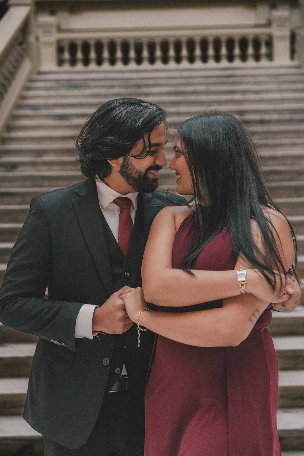 A couple dressed in formal attire, a man in a black suit and red tie and a woman in a red dress, sharing a close, intimate moment on a staircase.