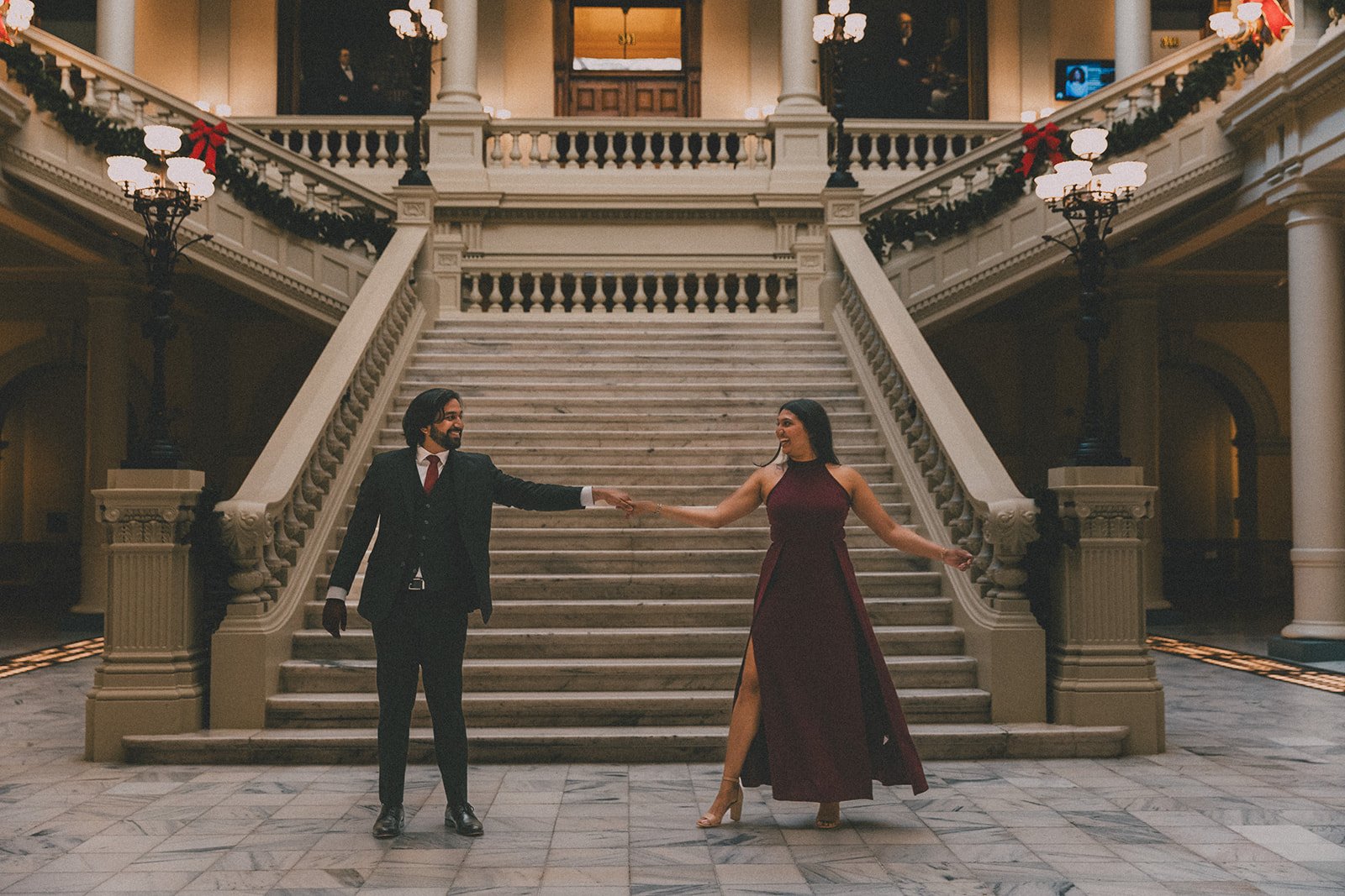 Two people, a man in a suit and a woman in a long dress, dancing and holding hands on a marble floor in front of a grand staircase decorated with Christmas garlands and lights in a historic building.
