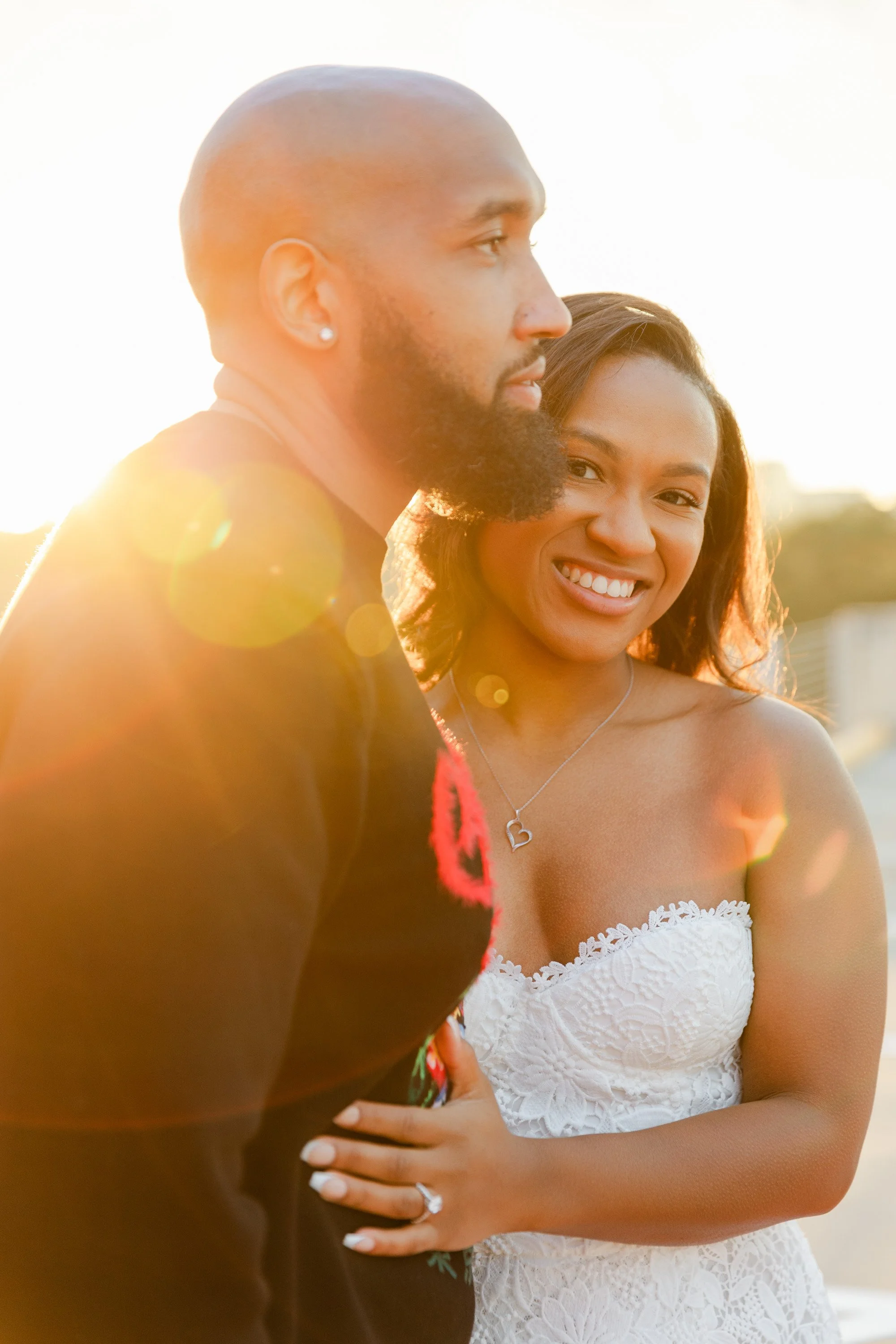 A smiling woman and a man celebrating outdoors at sunset, with the woman wearing a white strapless lace dress and the man in a dark shirt, the woman showing her engagement ring.