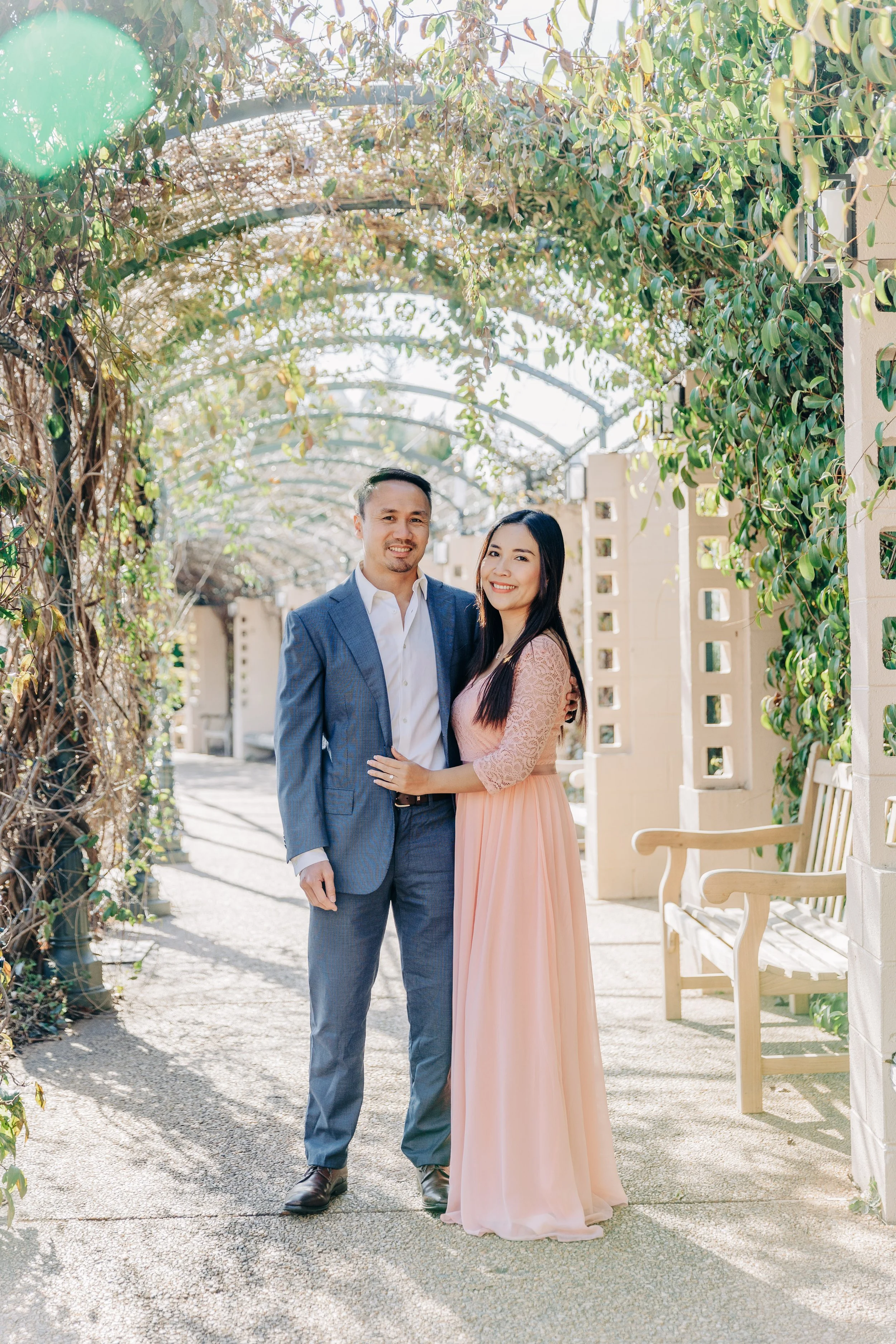 A couple outdoors on a sunny day, standing under a decorative archway covered with green foliage, smiling at the camera.