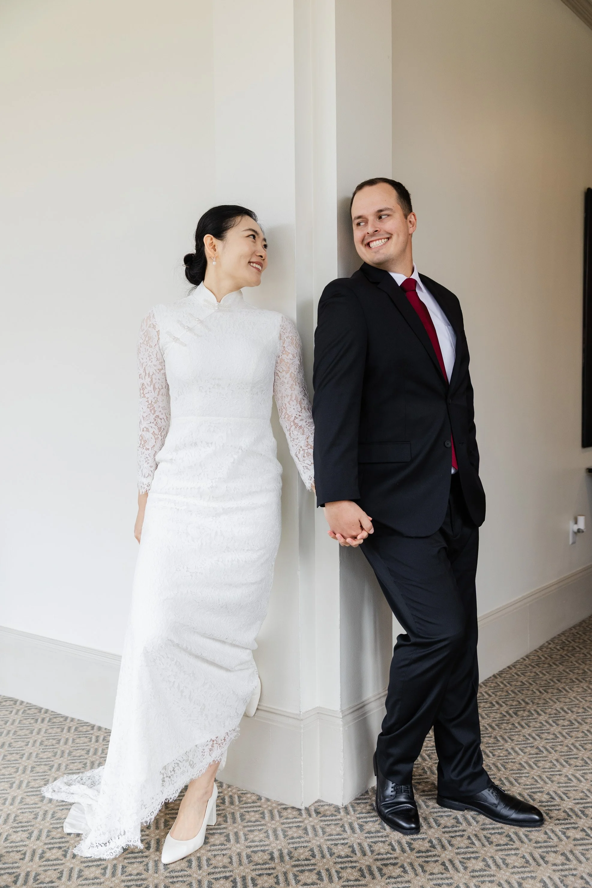 A bride and groom holding hands and smiling at each other, standing back-to-back against a wall in a room with a patterned carpet.