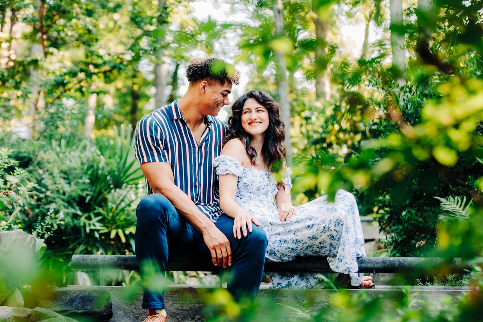 A young couple sitting on a park bench surrounded by green foliage, smiling and enjoying each other's company.