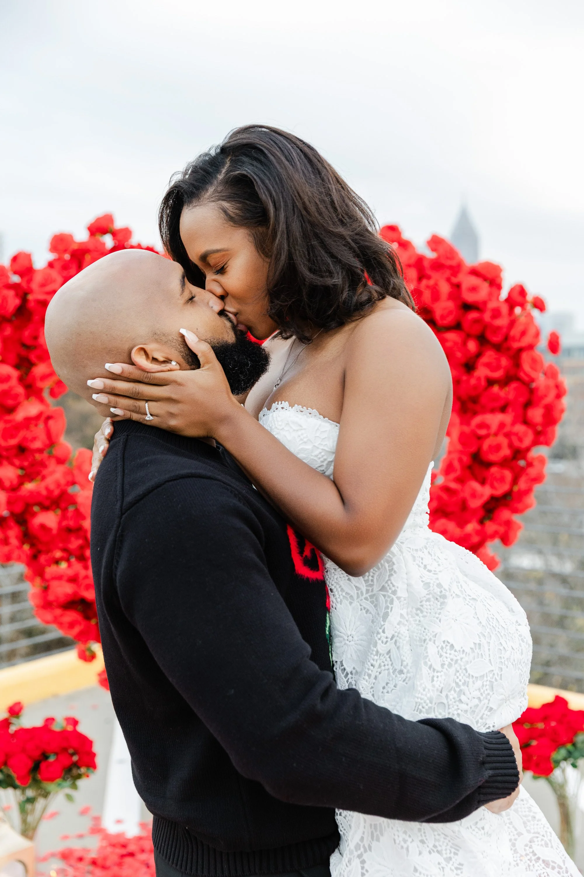 A couple sharing a romantic kiss in front of a large heart-shaped display of red roses on a rooftop.