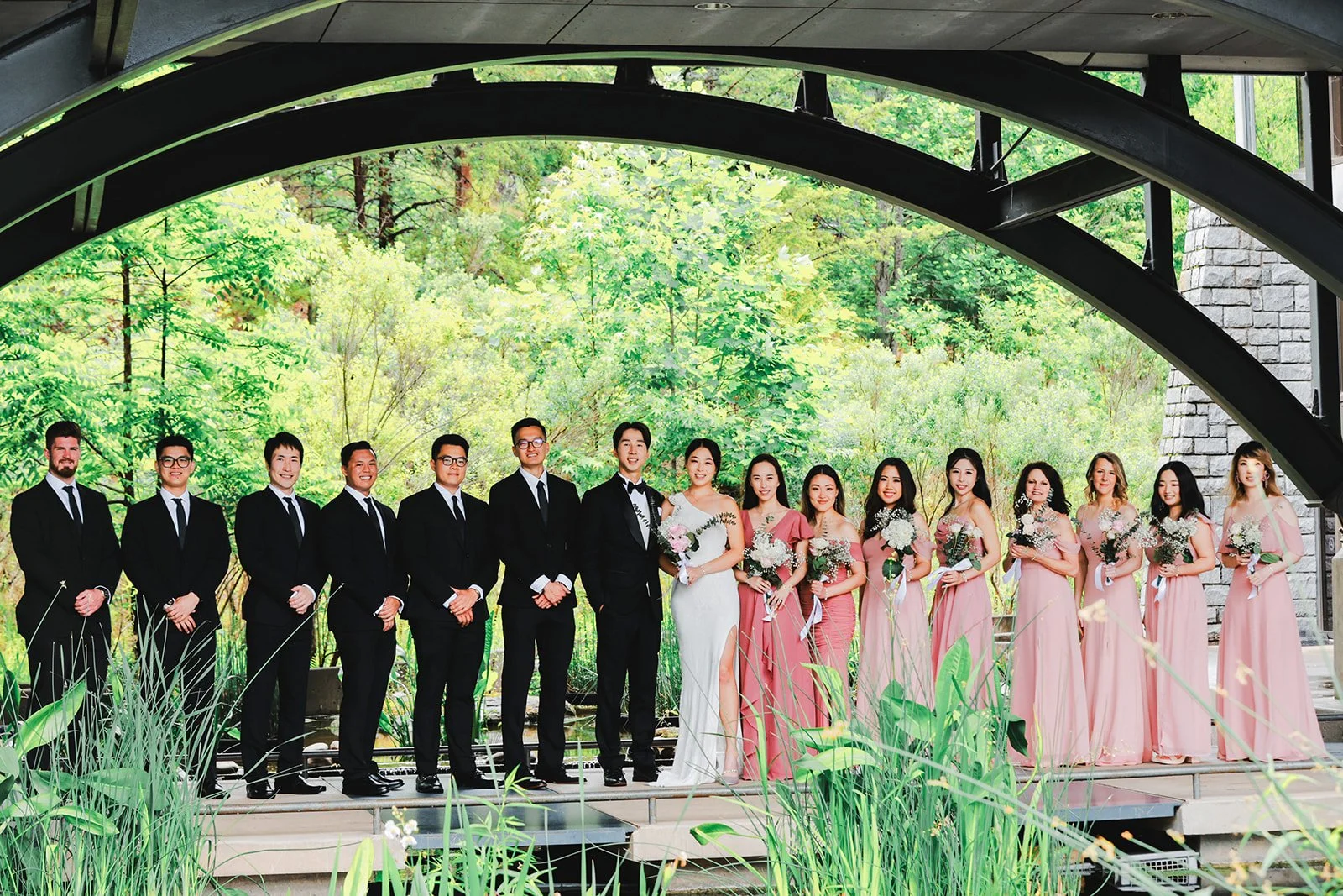 A wedding party standing on a wooden platform beneath a bridge with greenery in the background. The group includes the bride and groom, groomsmen in black suits, and bridesmaids in pink dresses holding bouquets.