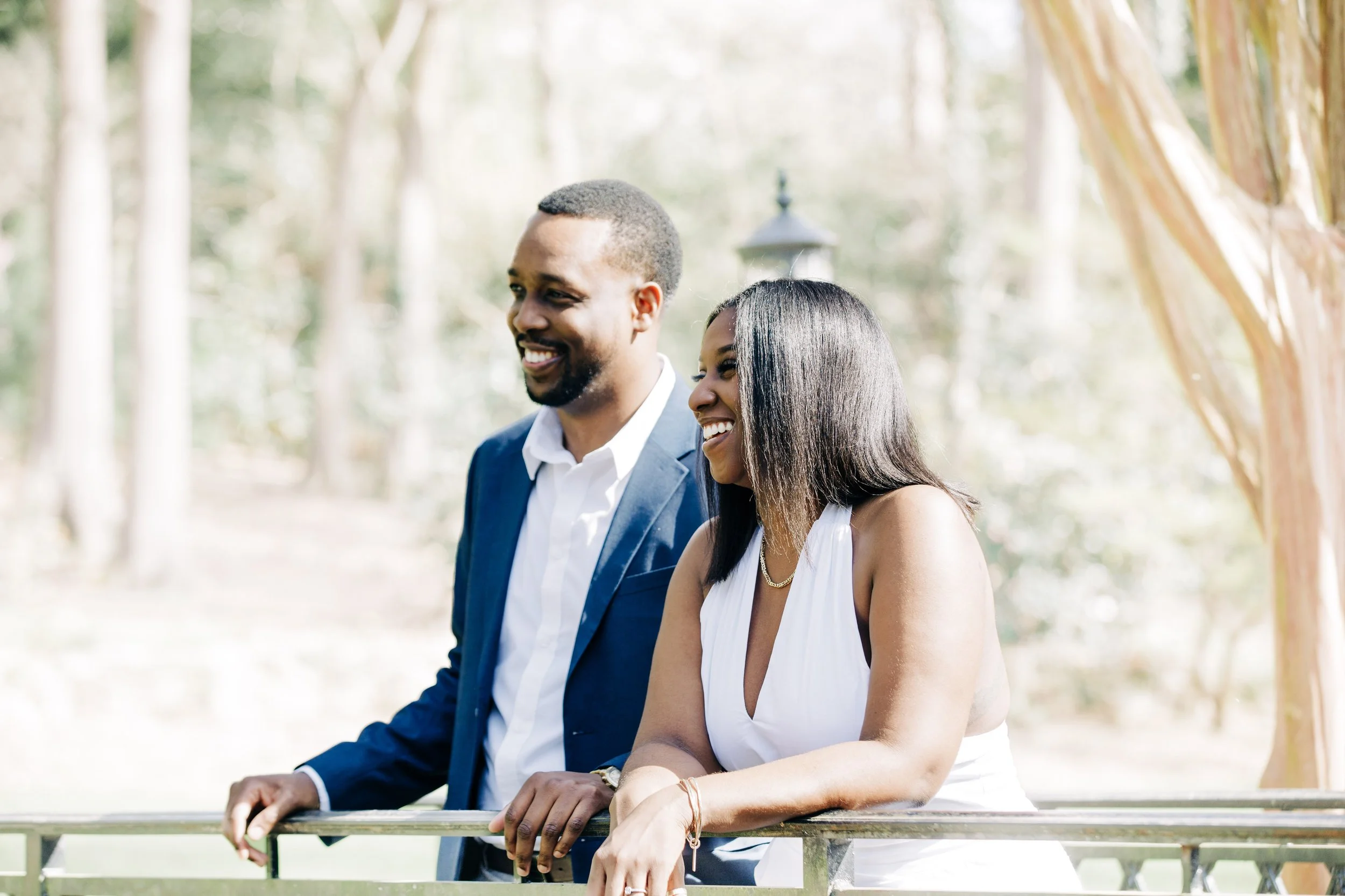 A smiling man in a blue suit and a woman in a white dress standing outdoors by a railing, with trees and sunlight in the background.