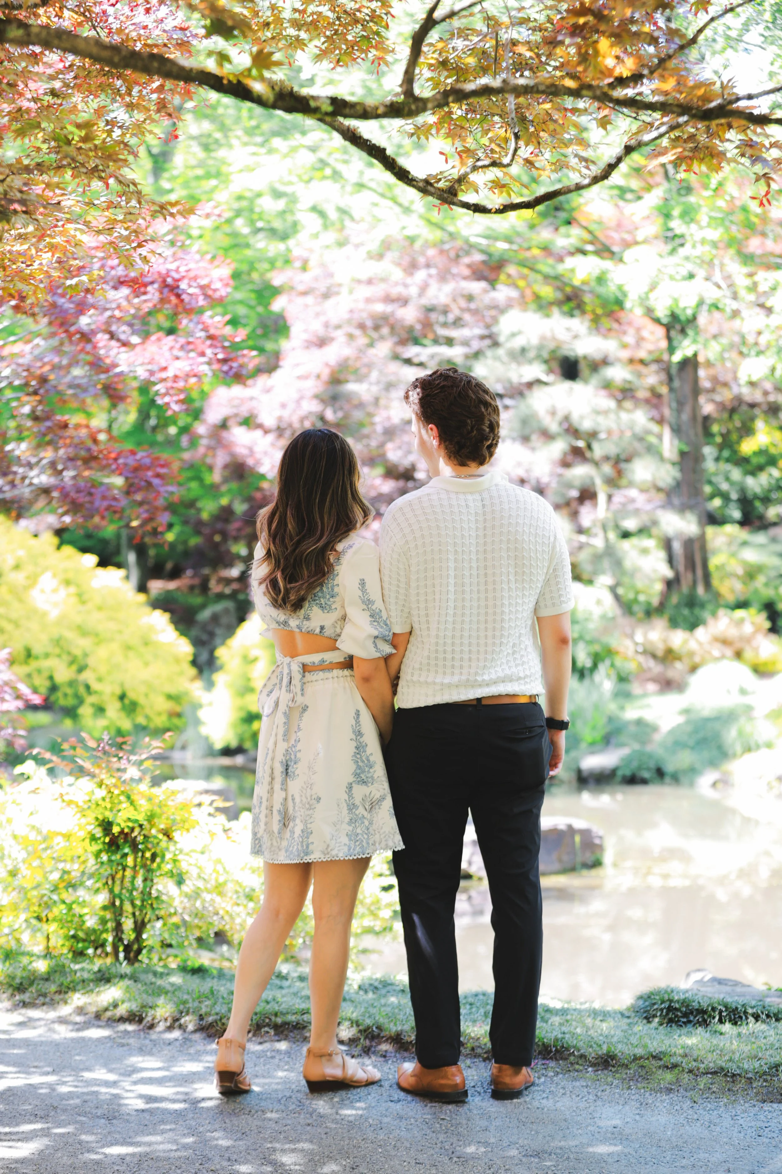 A couple walking in a colorful garden with trees and a small pond, viewed from behind.