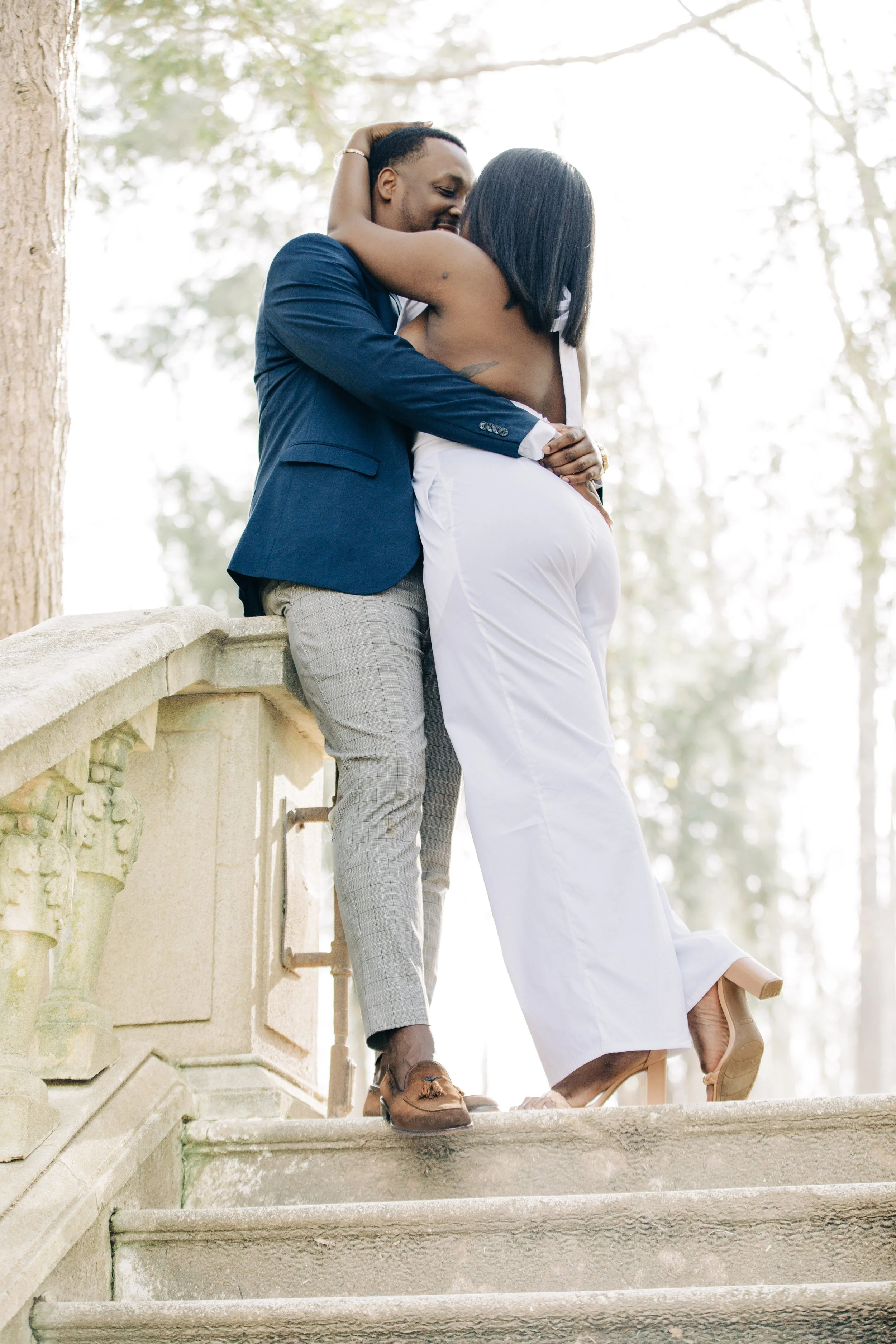 A couple hugging outdoors on stairs, with the man in a navy blazer and plaid trousers and the woman in a white dress, surrounded by trees.