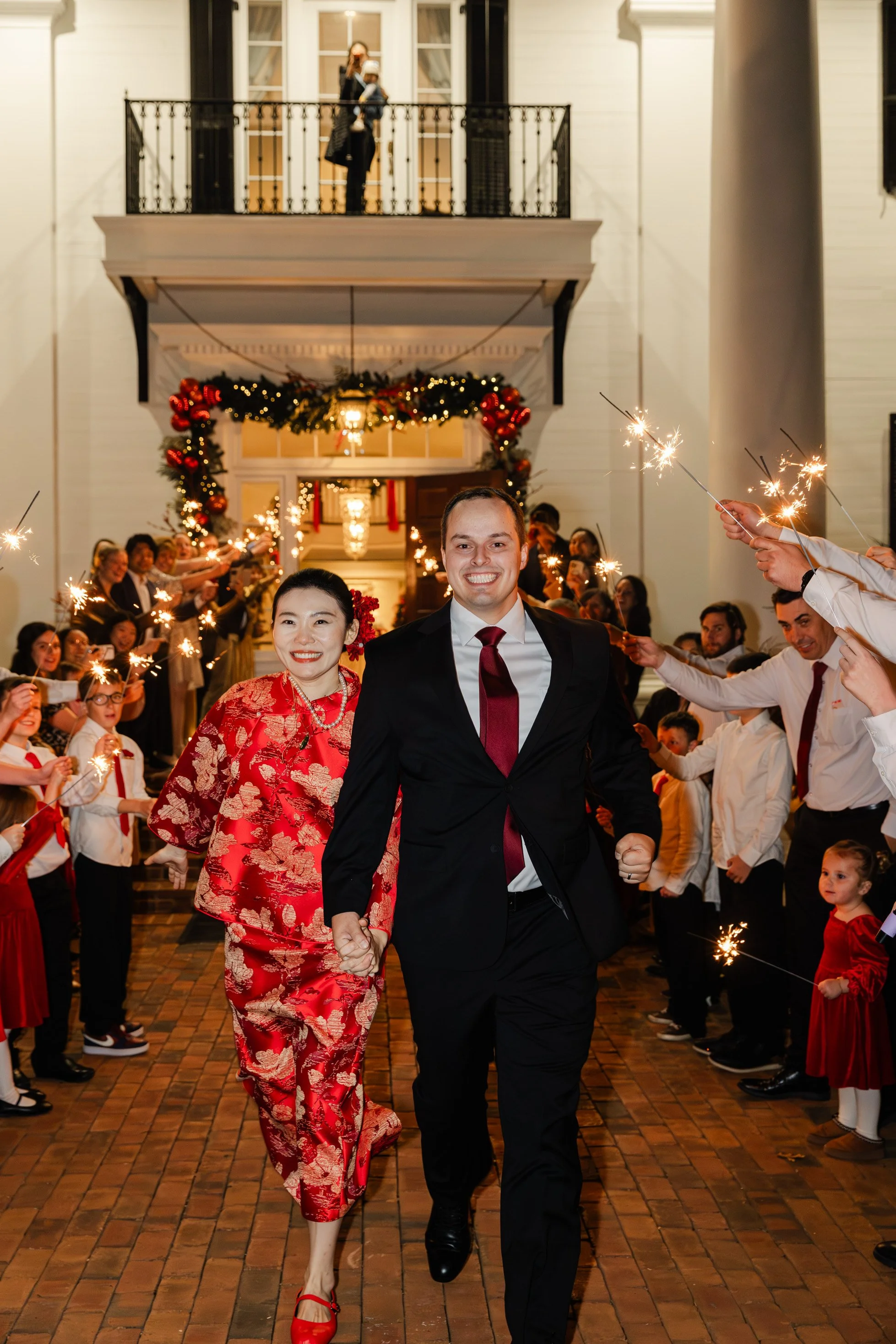A couple dressed formally, holding hands and smiling, is walking through a festive celebration with sparklers. The woman is in a red traditional dress, and the man is in a black suit with a maroon tie. The background shows an outdoor event decorated 