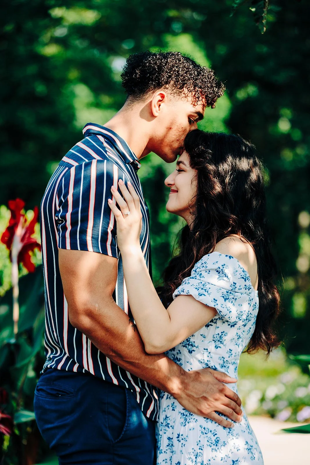 A couple in love sharing a tender moment outdoors, with the man kissing the woman's forehead as she smiles with closed eyes, surrounded by greenery.