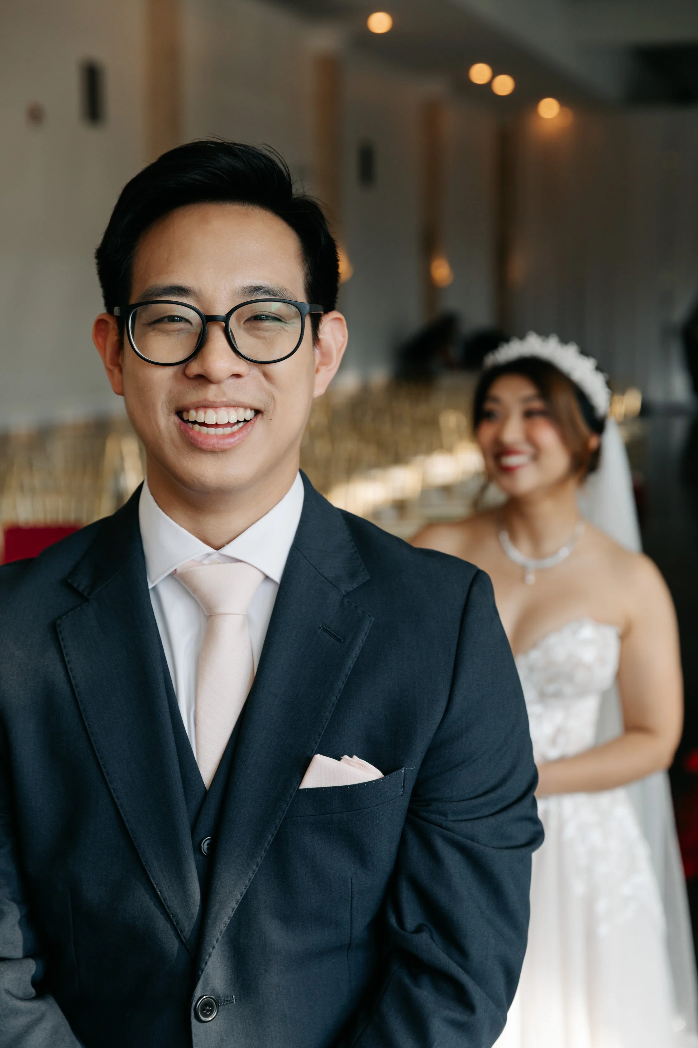 Smiling groom in black suit and glasses, with bride in white wedding dress and floral headpiece, in a decorated indoor venue.