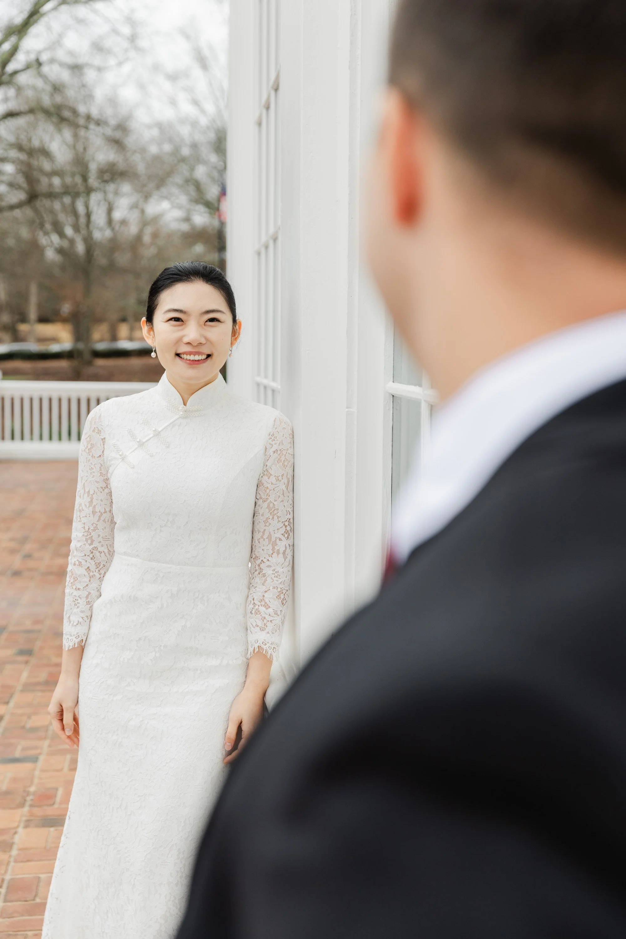 A woman in a white lace wedding dress smiling at a man in a black suit and red tie, outside near white building and brick pavement.