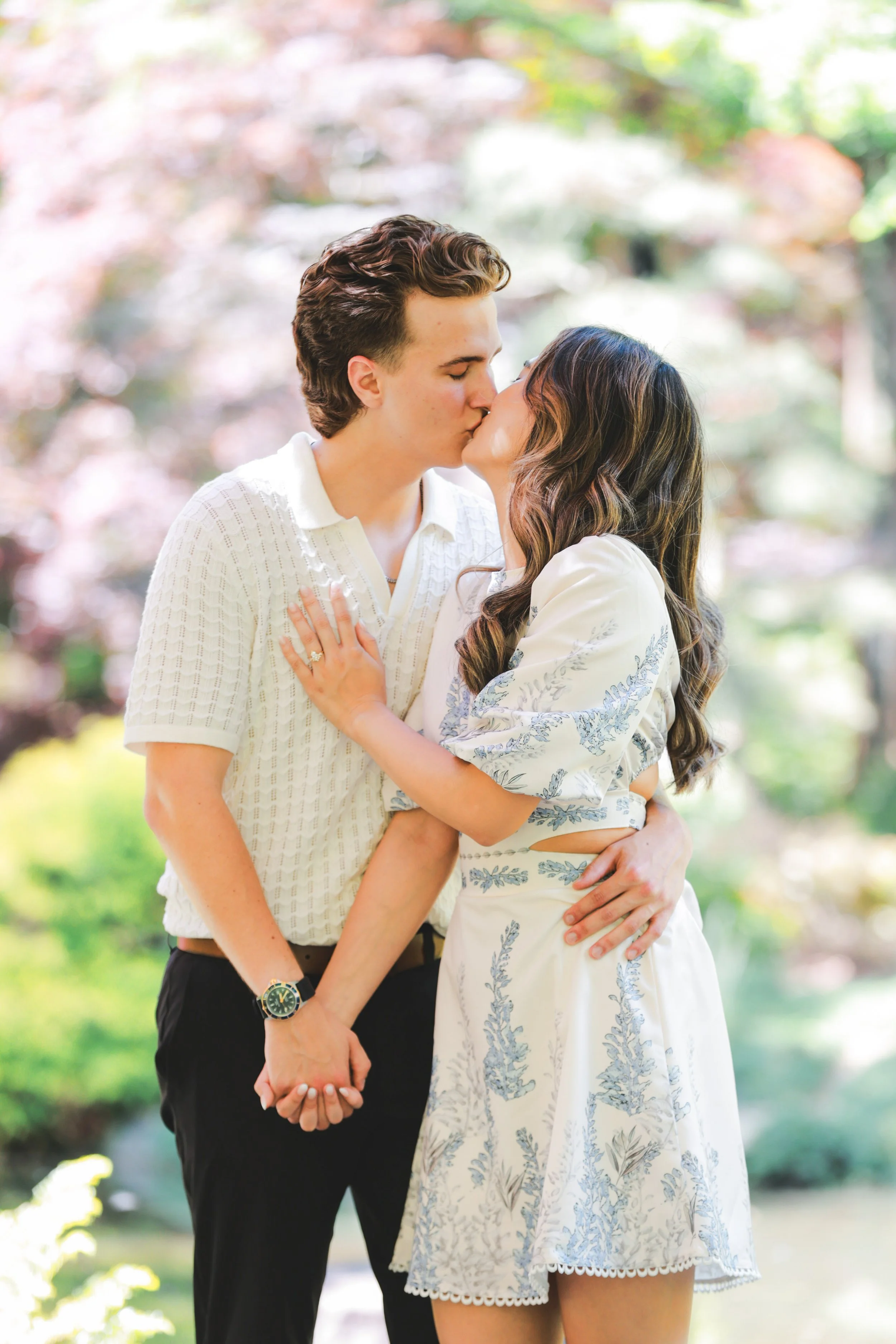 A couple sharing a kiss outdoors, holding hands, surrounded by greenery and colorful trees.