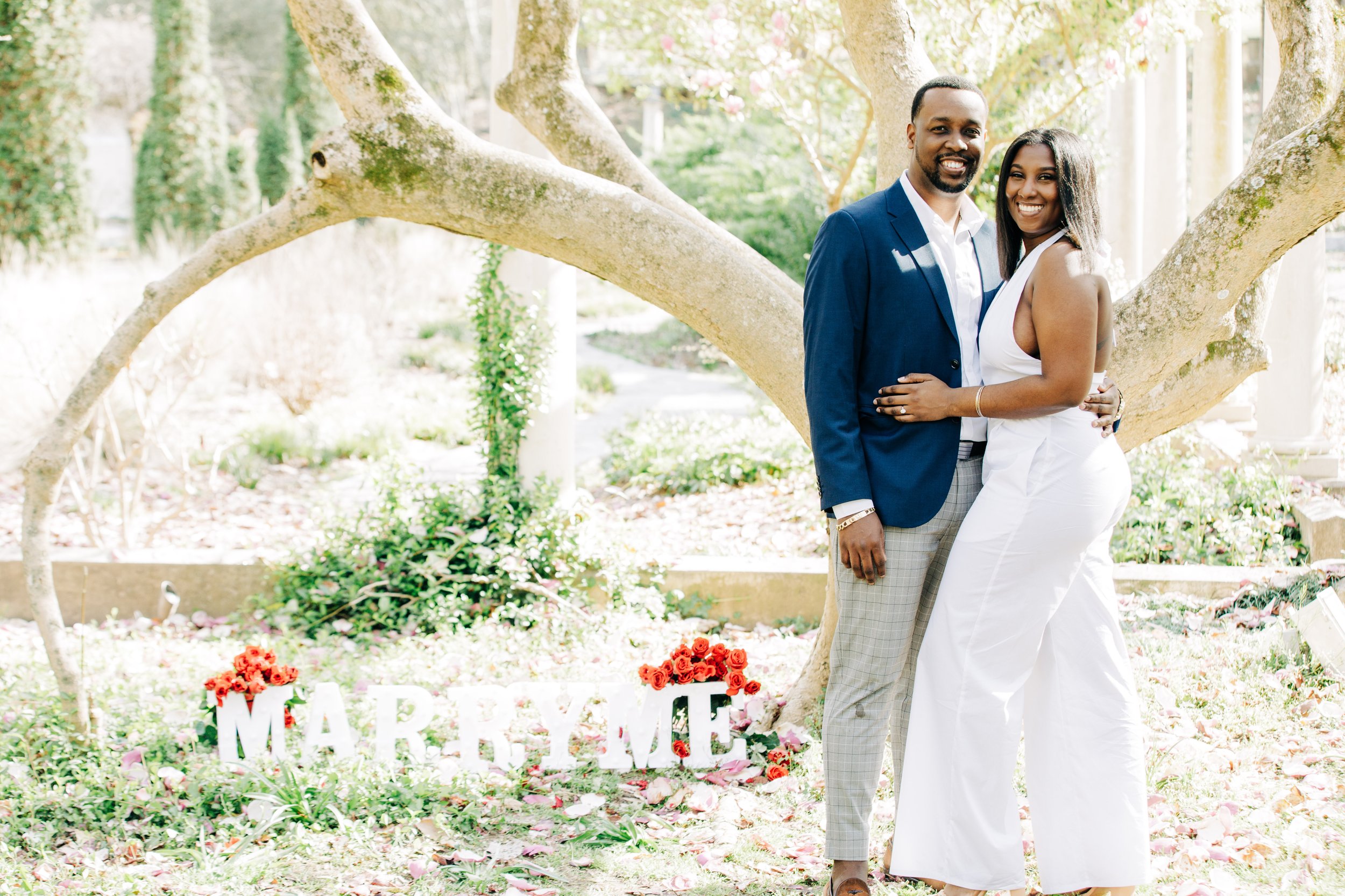 An African American couple is smiling and embracing under a large tree in a garden, with a sign that spells 'MARRY ME' decorated with red flowers at their feet.