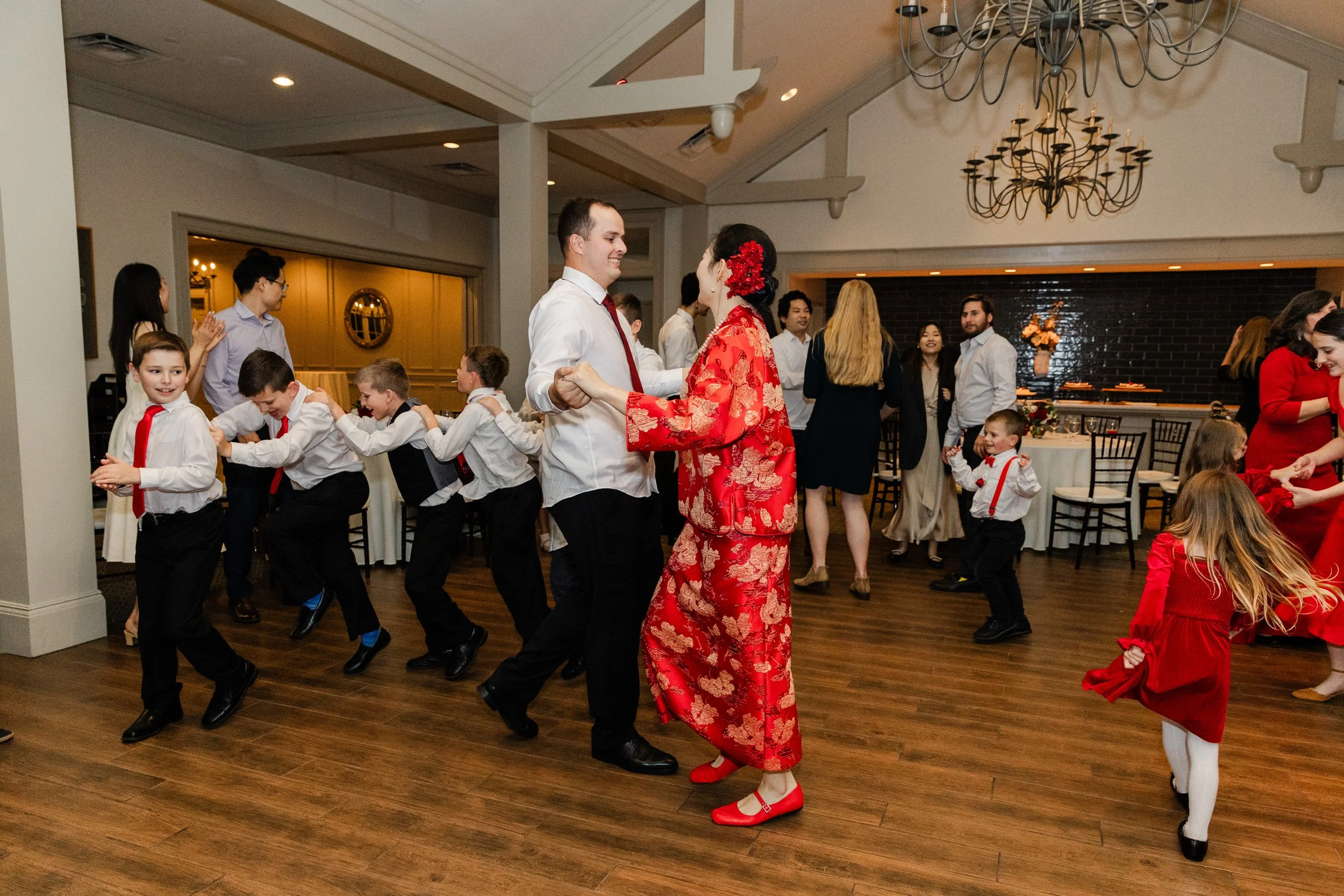 People dancing and playing at a party or celebration, including children and adults, with some children in a conga line and others dancing with partners in a decorated indoor venue.