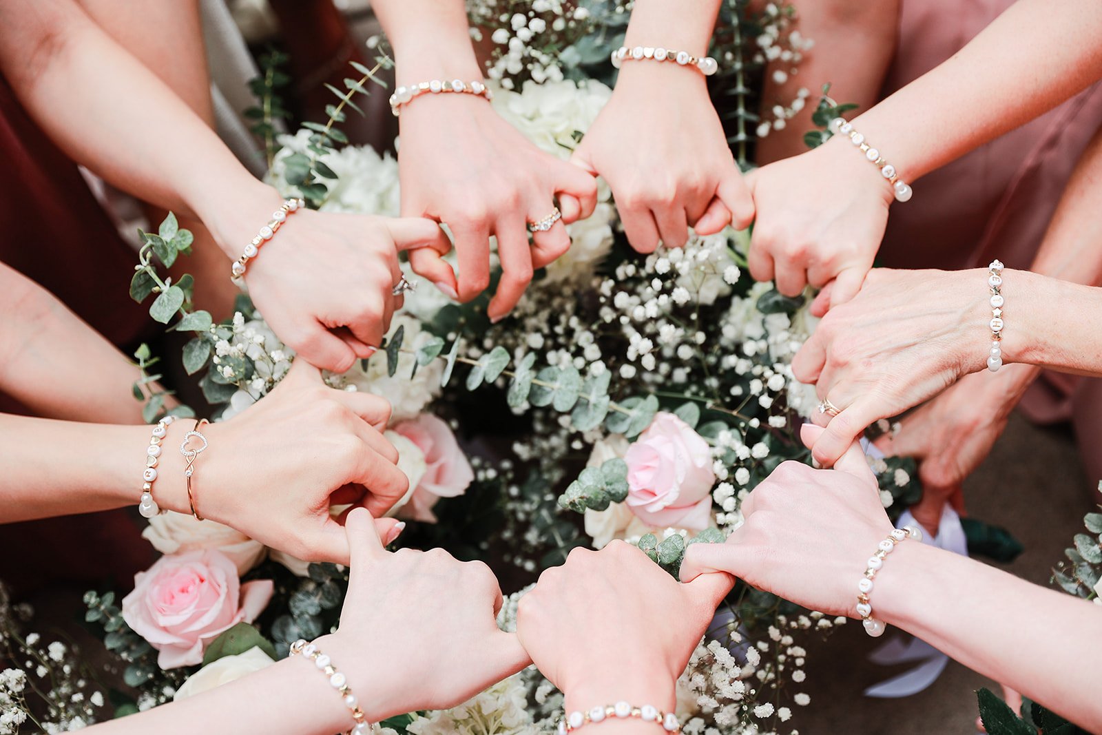 Multiple women’s hands forming a heart shape over a floral arrangement of pink roses and white baby's breath, all wearing pearl jewelry.