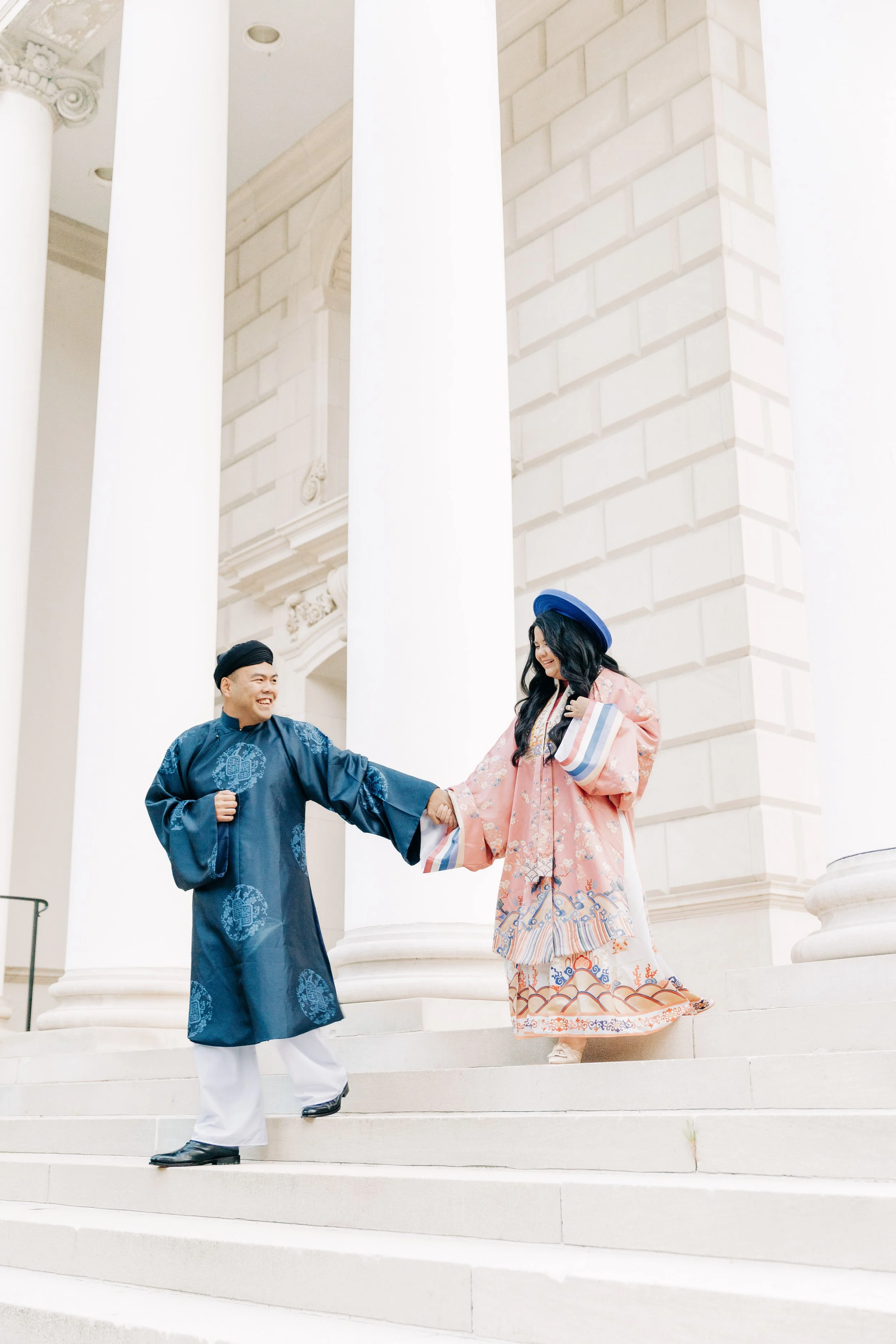 A couple dressed in traditional Korean clothing, Hanbok, on the steps of a building with large white columns, smiling and holding hands.